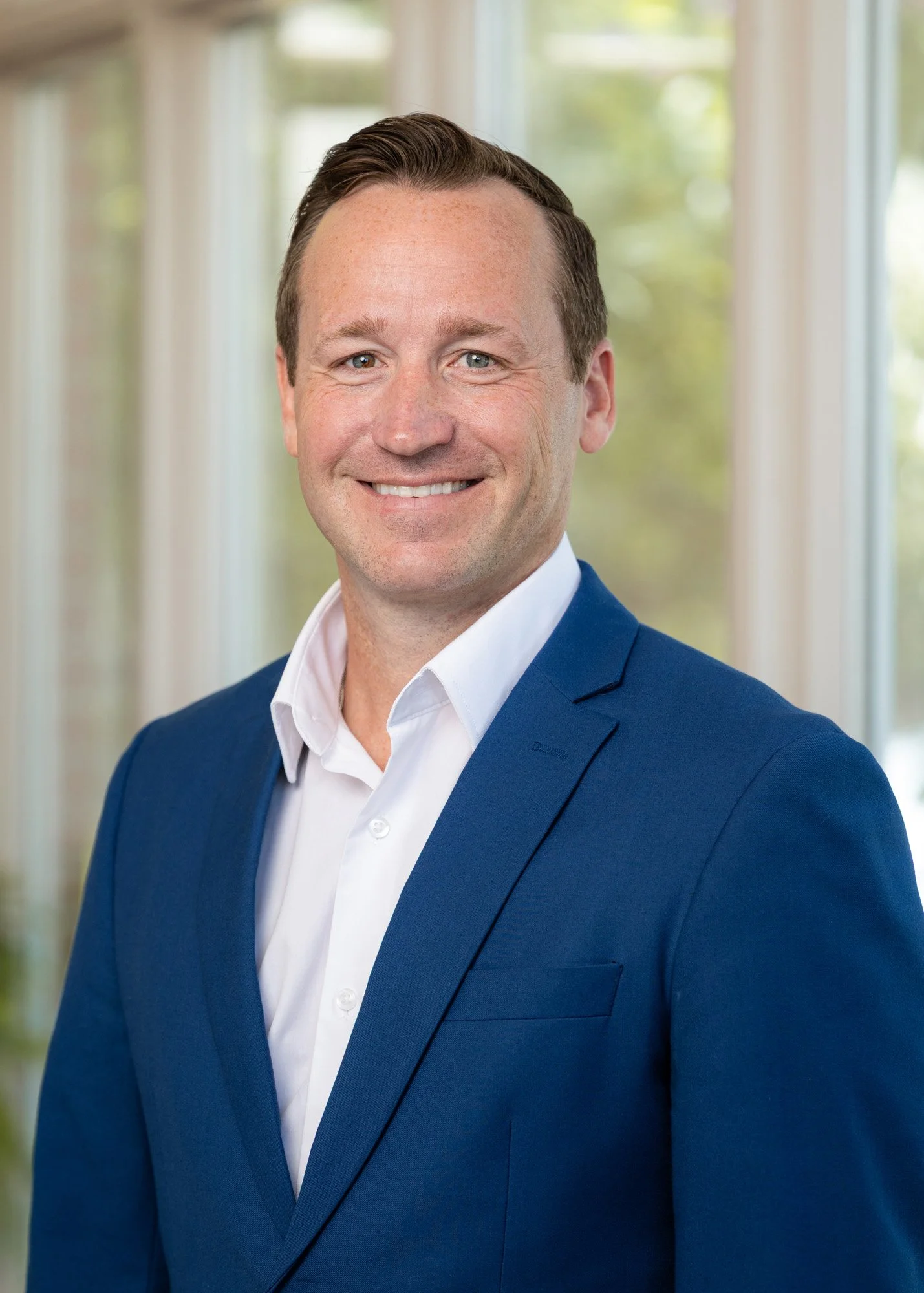 A portrait of a smiling man dressed in a blue suit and white shirt, with a blurred bright background of windows and greenery.