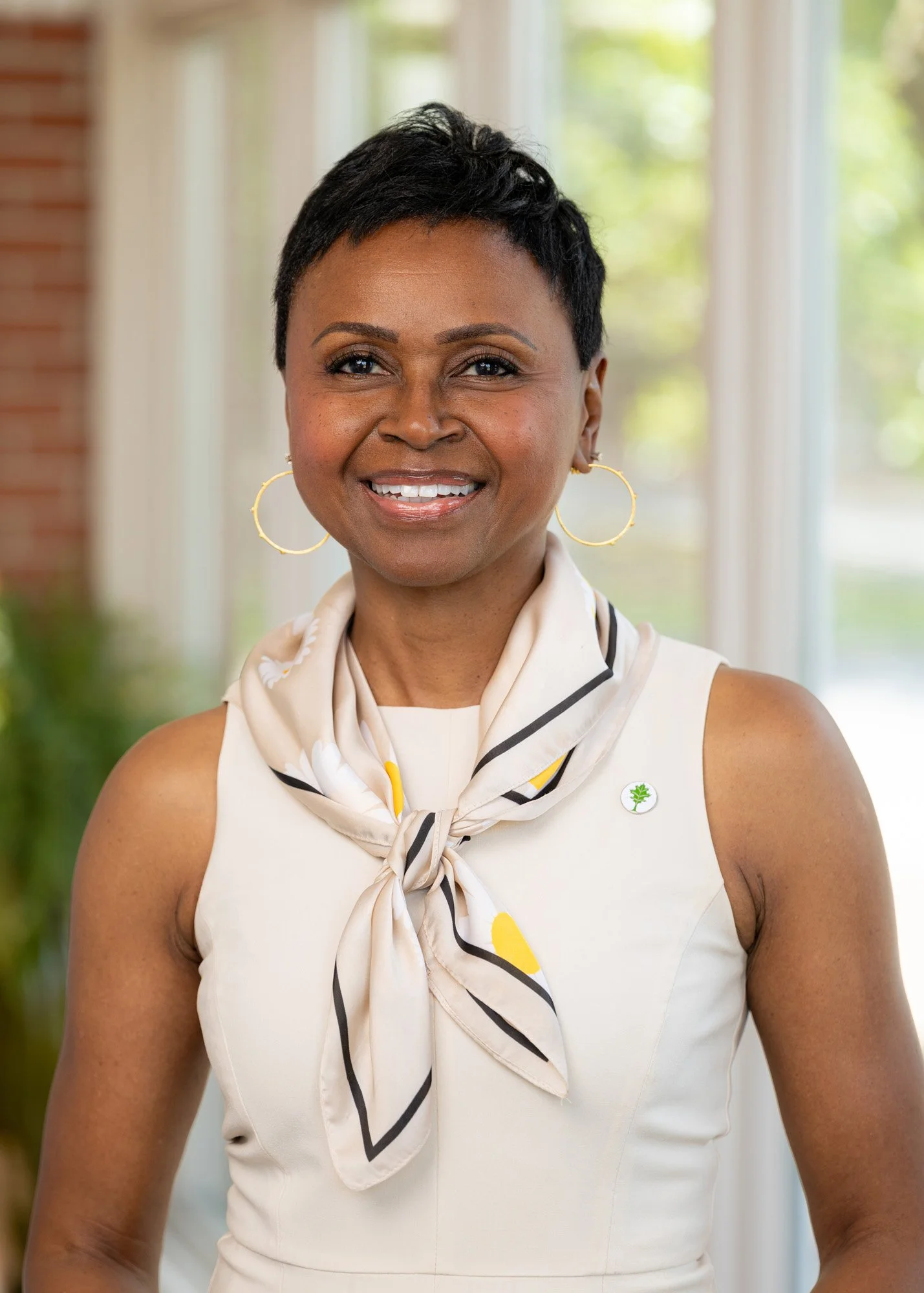A woman with short black hair wearing gold hoop earrings and a sleeveless white top with a beige scarf tied around her neck, standing indoors with a blurred green outdoor background visible through large windows.