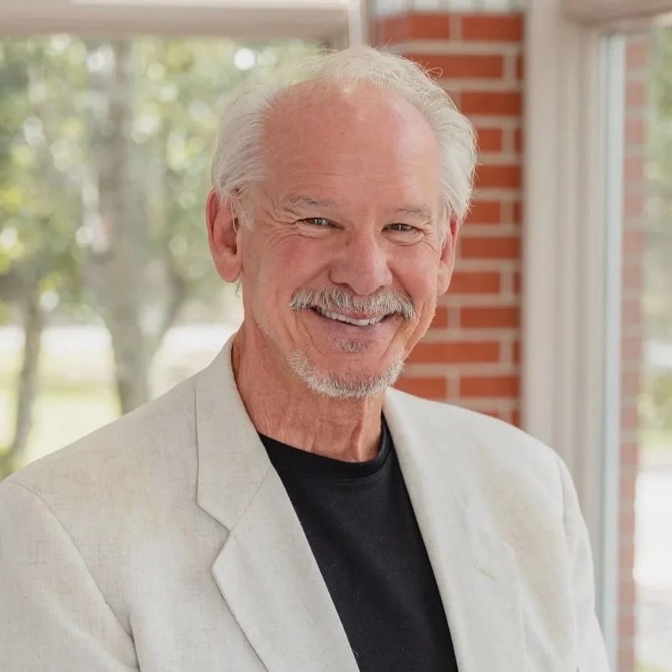 A smiling elderly man with white hair, a mustache, and a beard, wearing a light-colored blazer over a black shirt, standing indoors near a window with trees outside and brick wall in the background.