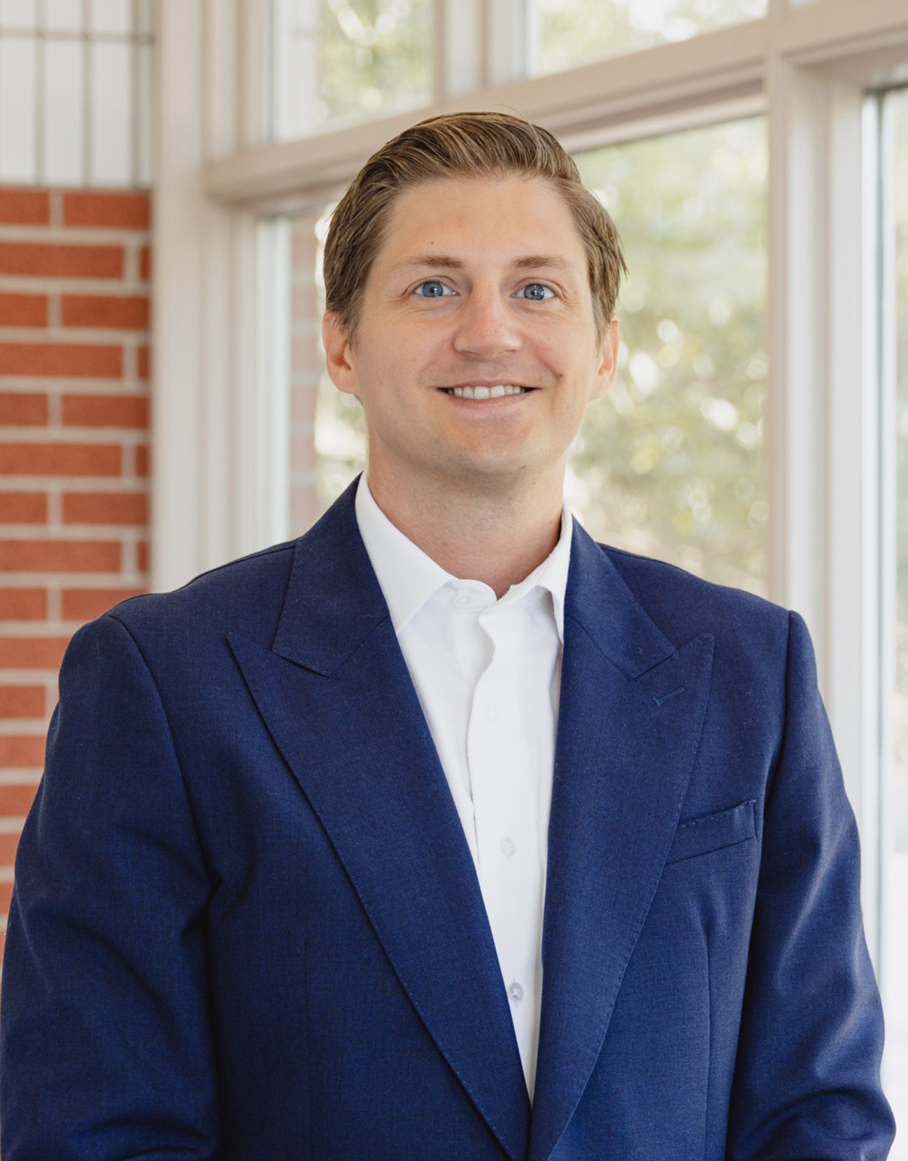 A smiling man in a dark blue suit and white shirt standing indoors near a window with trees outside.