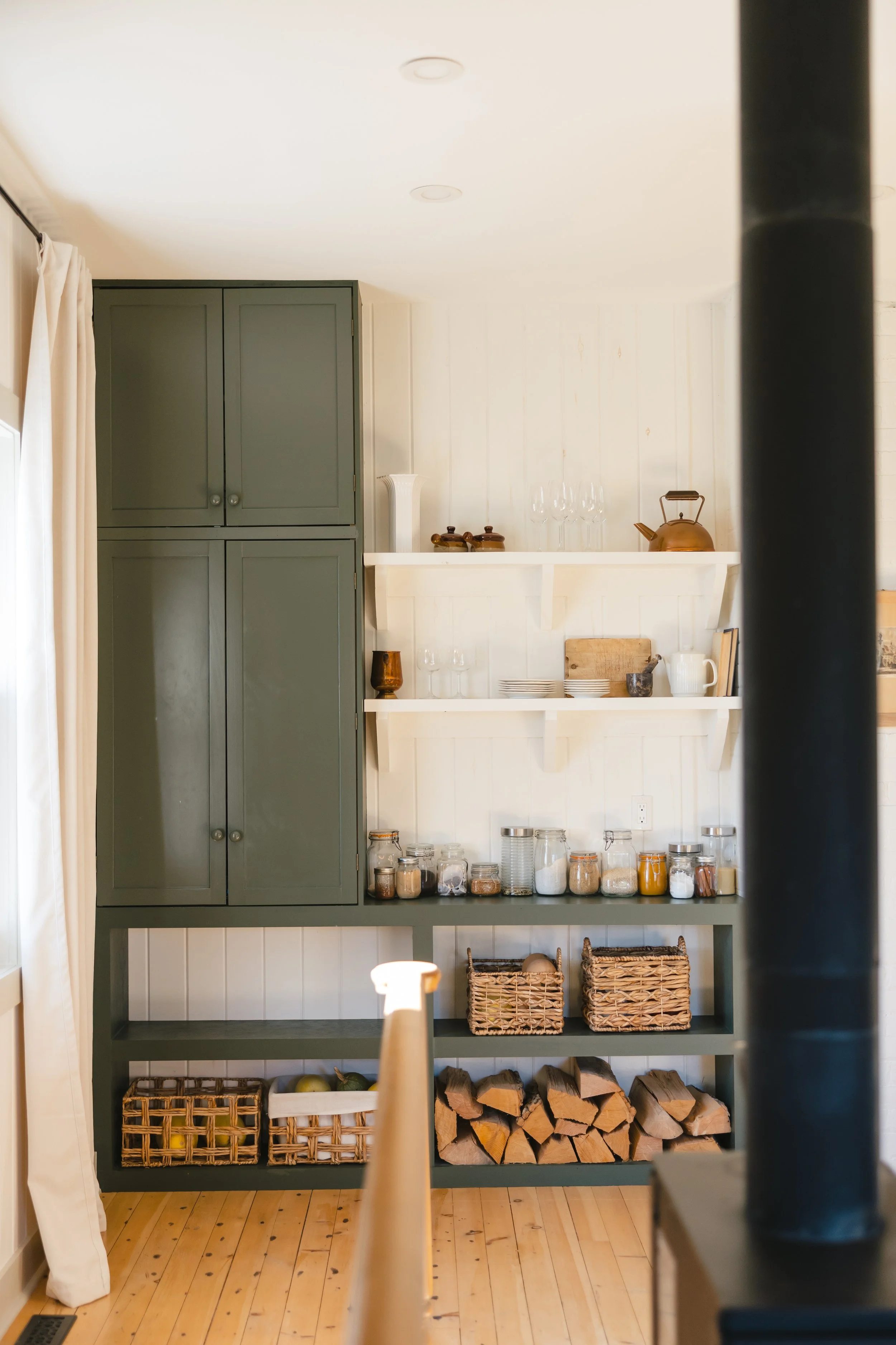 A view of a kitchen cabinet with open shelves displaying various glass jars, ceramics, and a teapot, with wood logs stored underneath on a wooden floor.