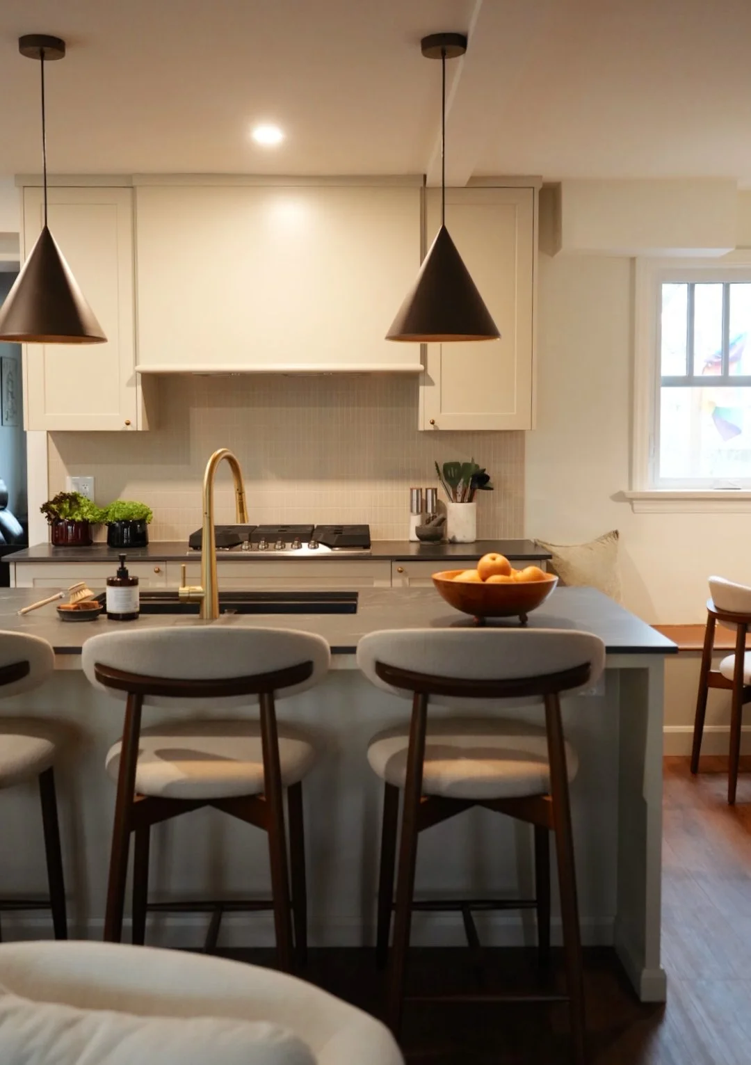 Modern kitchen with a central island, bar stools, pendant lights, and a window, featuring a bowl of oranges on the counter.