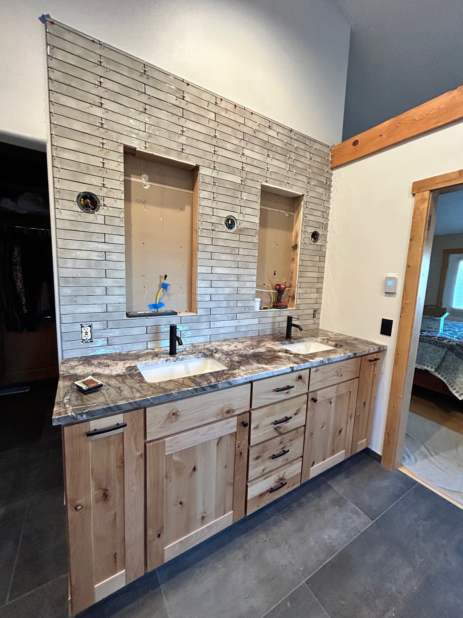 Bathroom vanity with dual sinks, marble countertop, wooden cabinets, and a partially completed tile wall behind for a mirror installation.