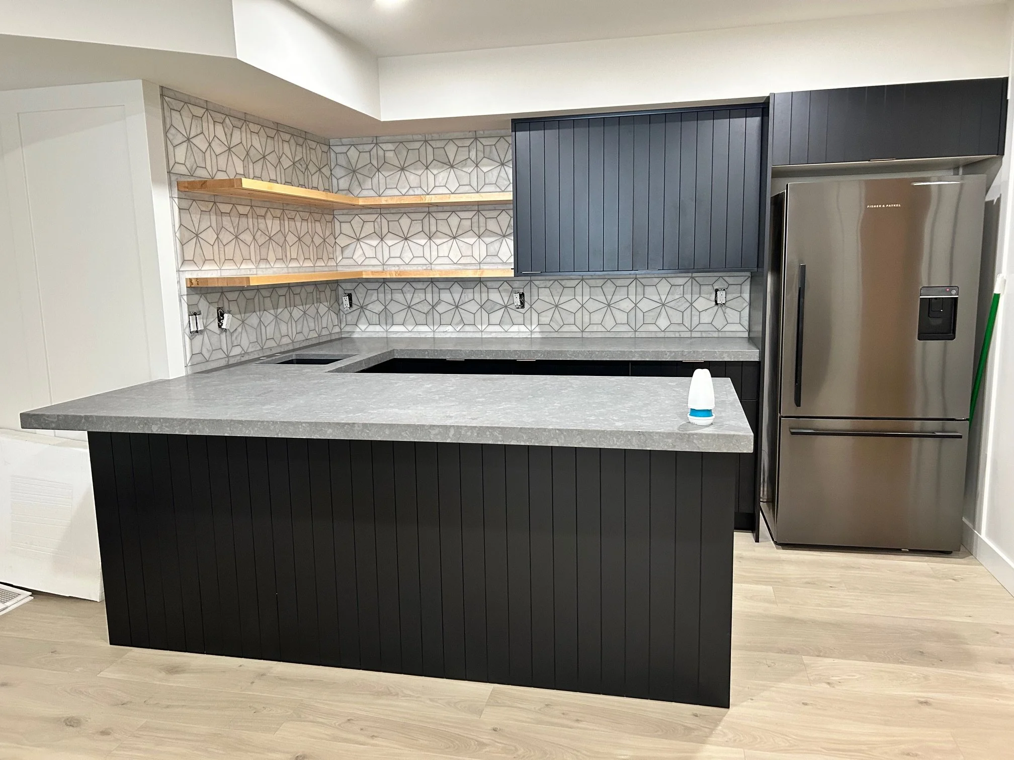 Modern kitchen with black cabinets, open wooden shelves, patterned tile backsplash, gray countertop, stainless steel refrigerator, and a small white object on the counter.