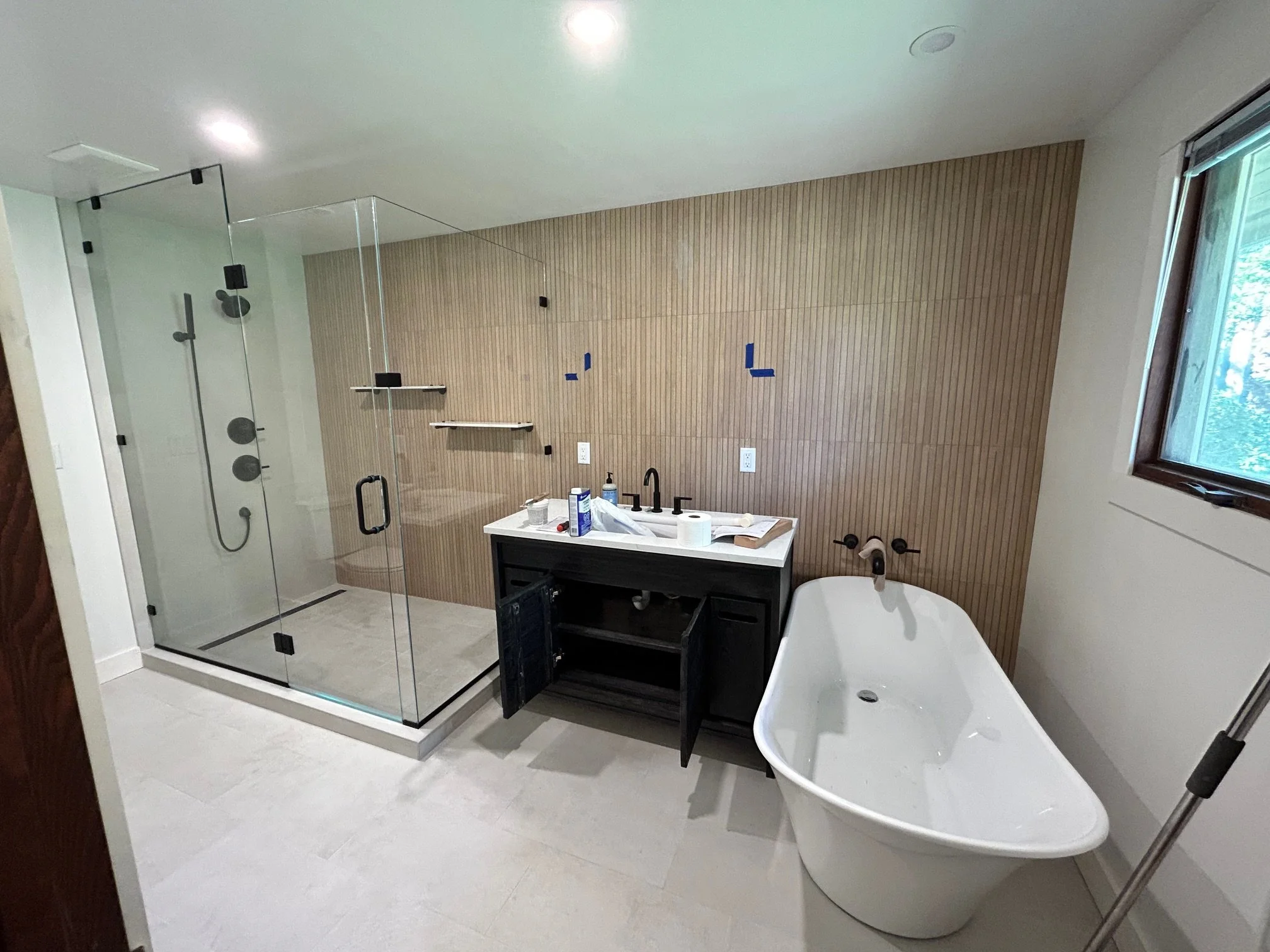 Modern bathroom with a glass-enclosed shower, black vanity with sink, and freestanding bathtub near a window, with wooden panel wall and light-colored floor tiles.
