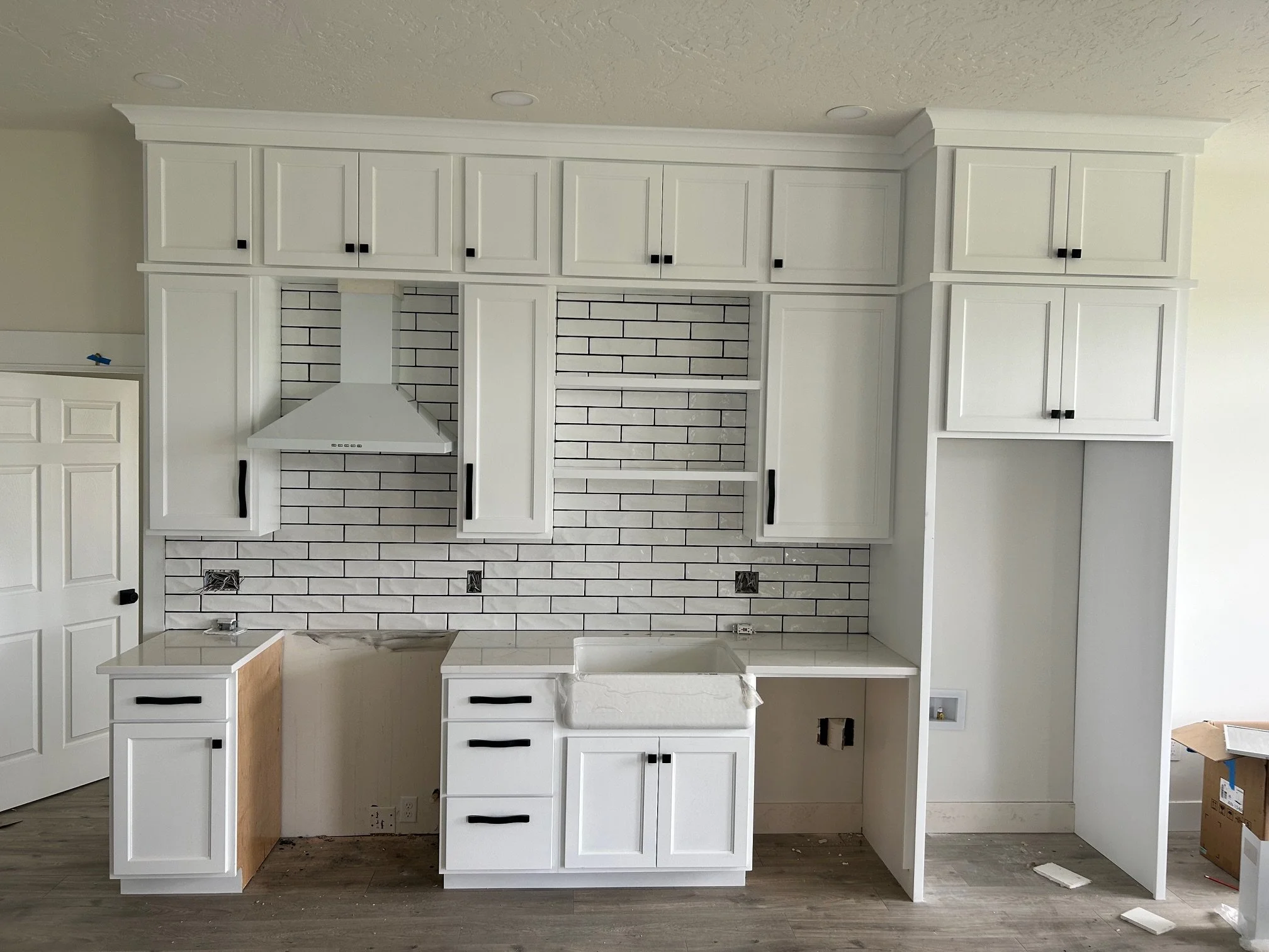 Kitchen cabinets under construction with white paint, black handles, black and white subway tile backsplash, a stove hood, and an unfinished countertop area.