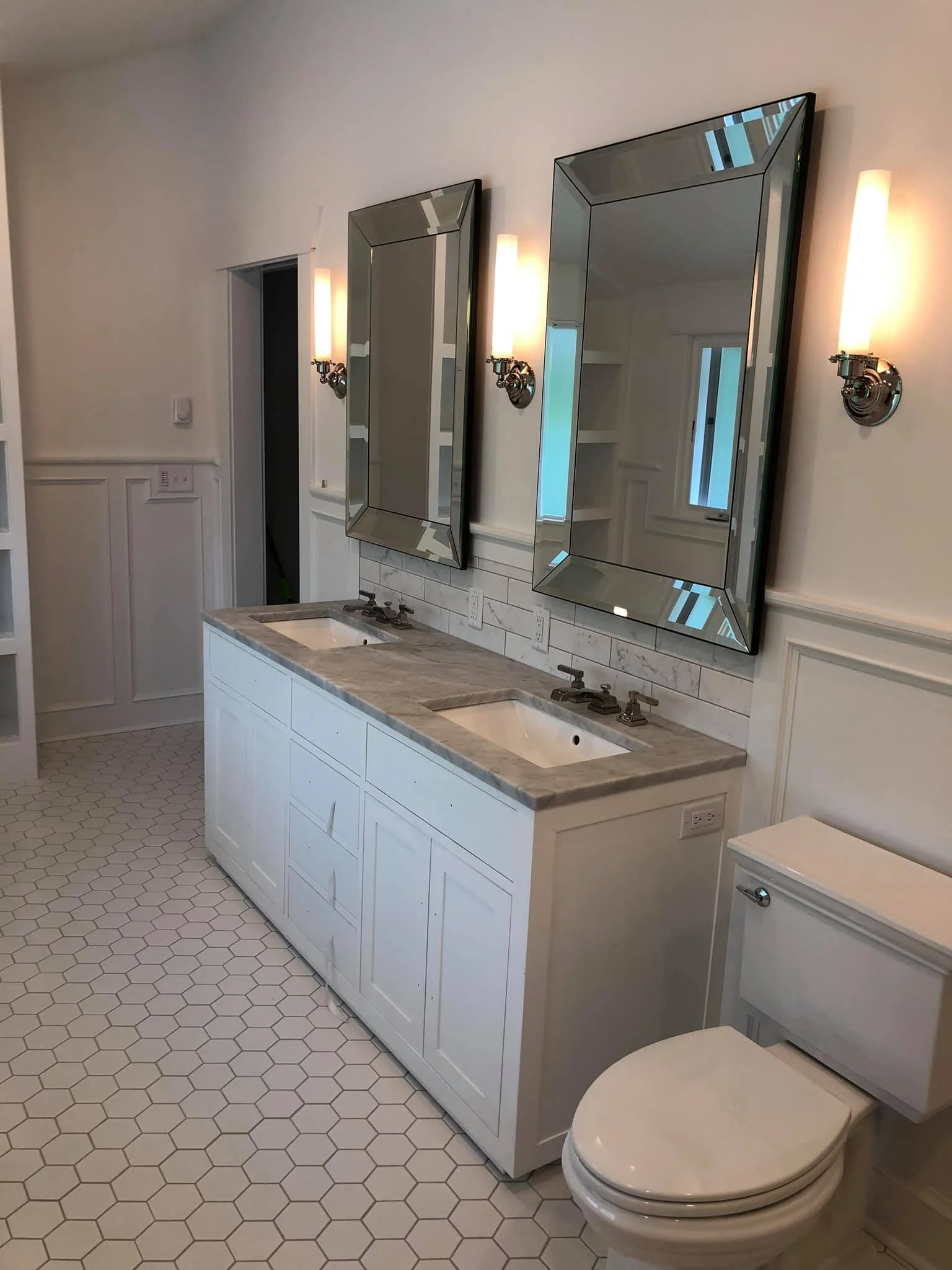 A bathroom with a double vanity with a marble countertop, two mirrors, two wall-mounted light fixtures, a toilet, and hexagonal floor tiles.