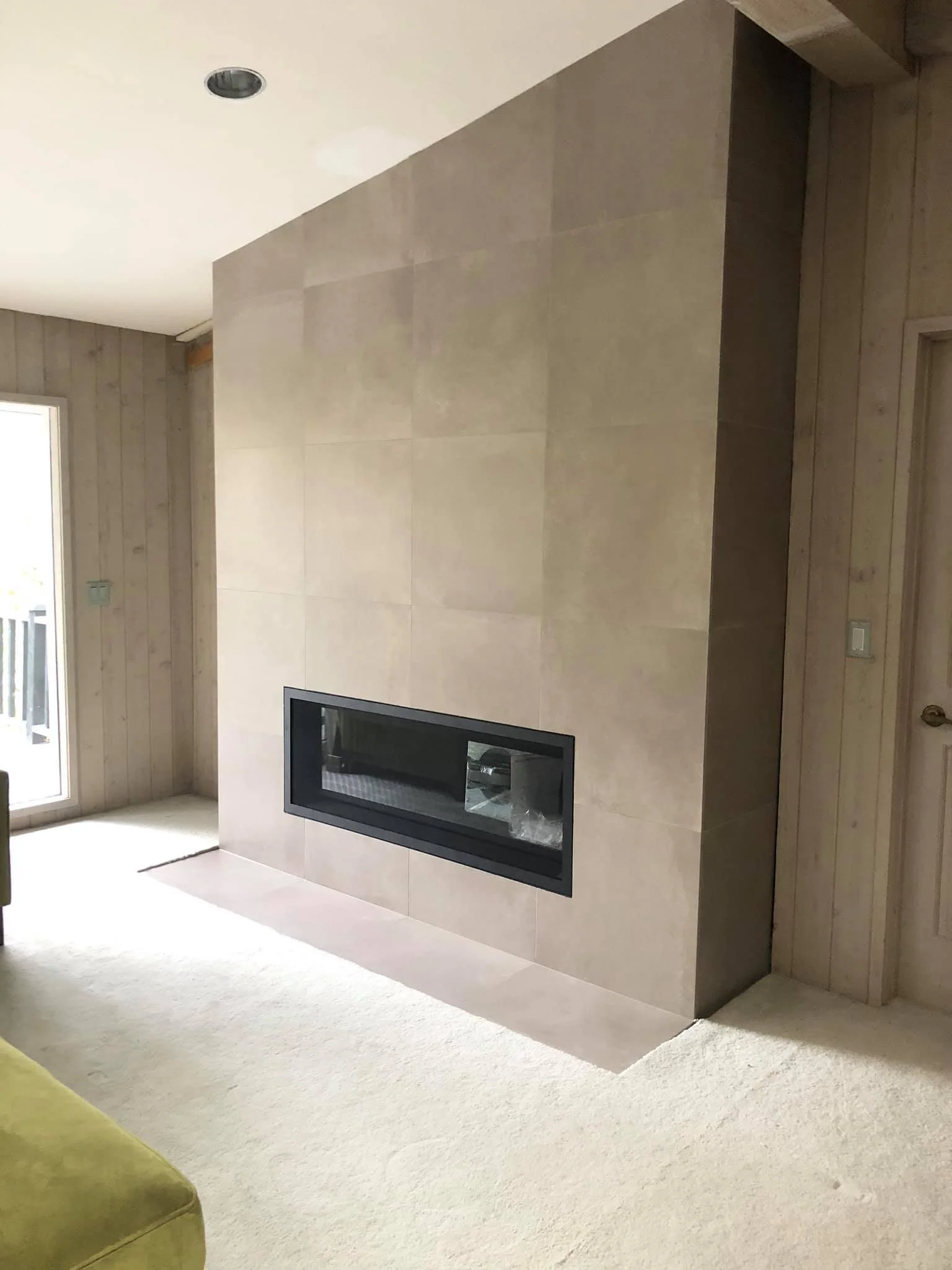 Modern living room fireplace with black-framed glass door set into a beige tiled wall, adjacent to wood-paneled walls and a sliding door leading outside.
