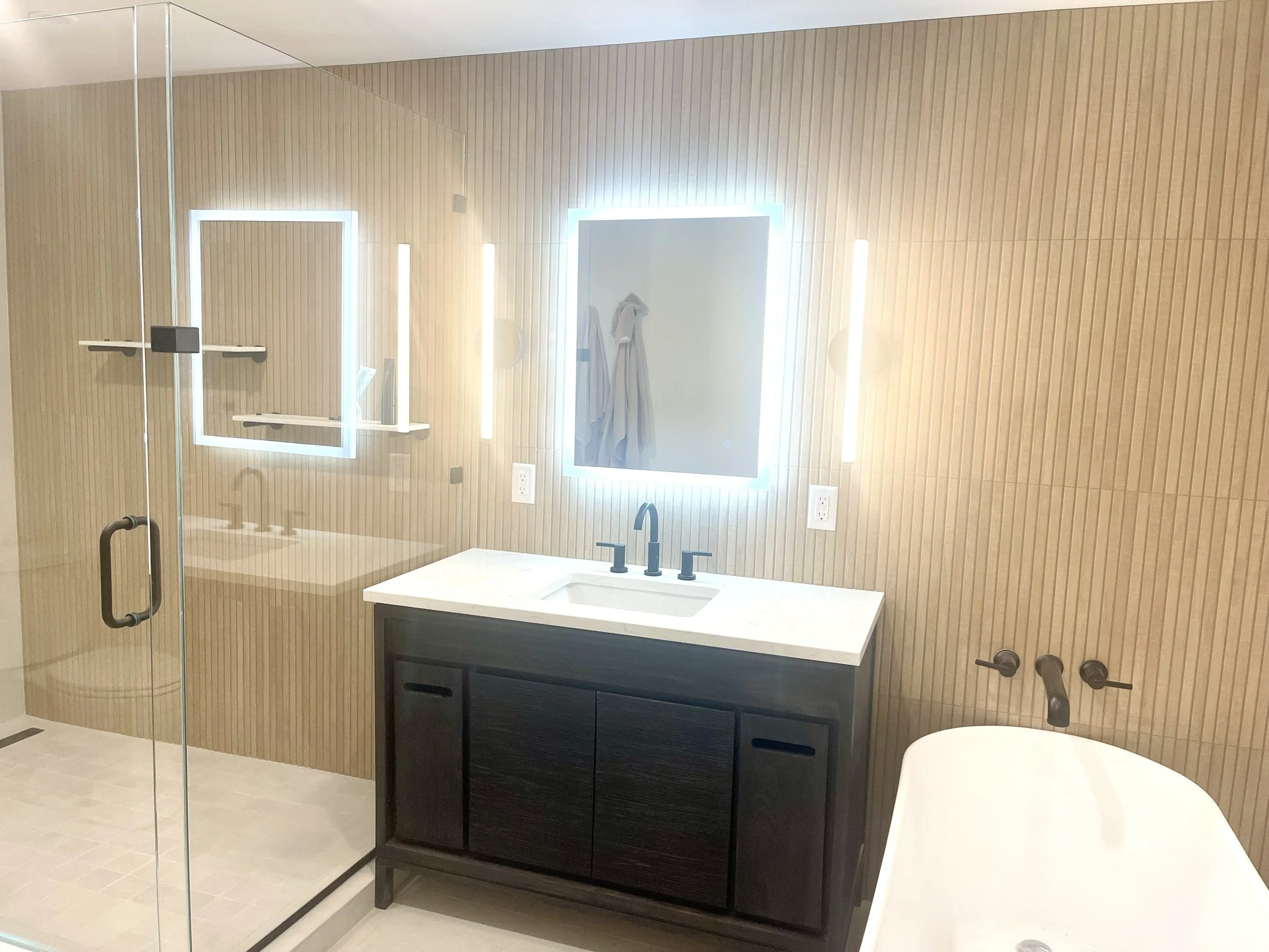 Modern bathroom with a glass shower enclosure, a black vanity with a white countertop and a rectangular mirror with backlit LED lighting, and a white bathtub with black fixtures, all against a wood-paneled wall.