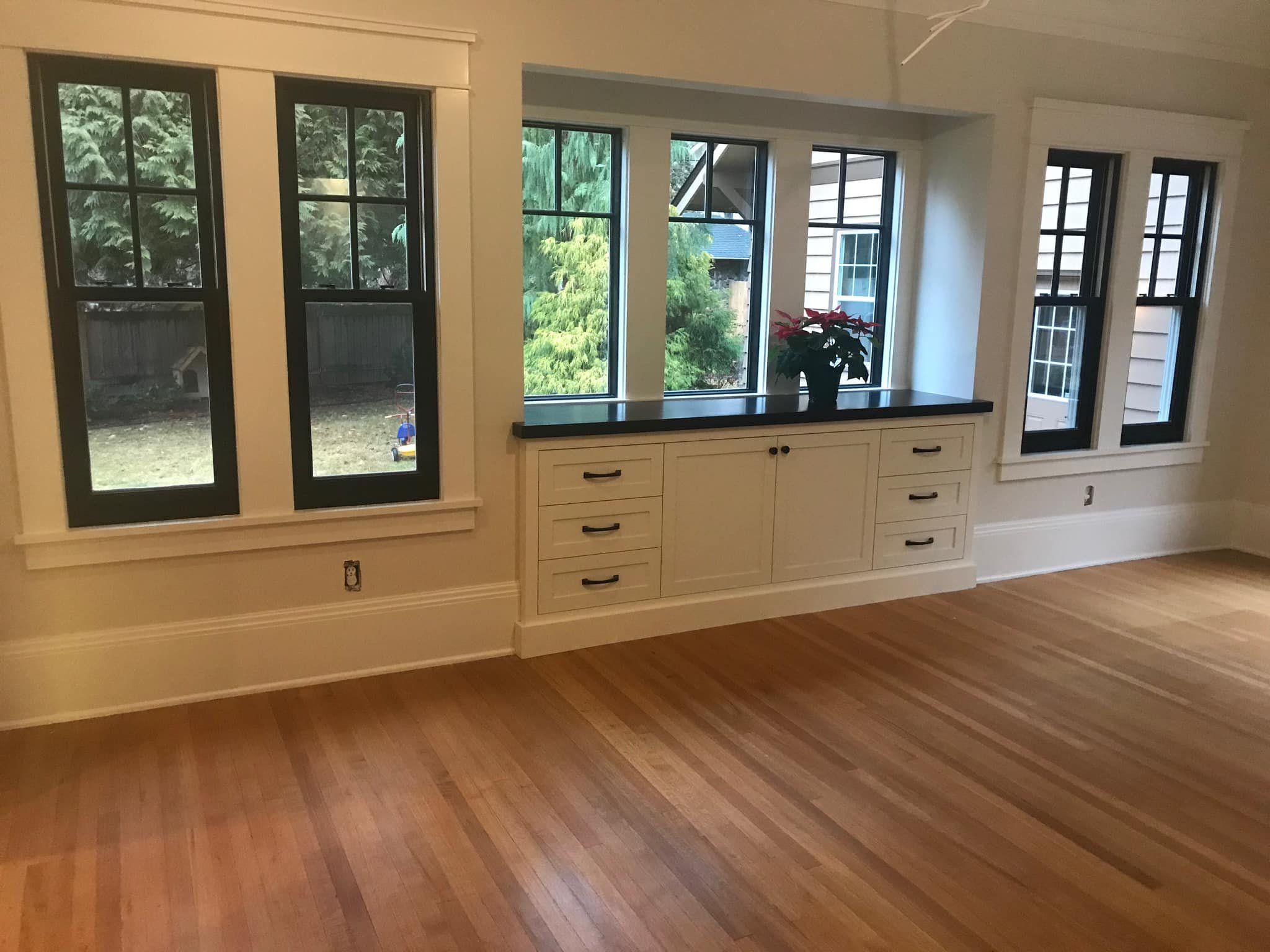Empty room with hardwood floors and multiple large windows with black trim, white walls, a built-in white cabinet with black handles and a black countertop, and a potted plant on the cabinet.
