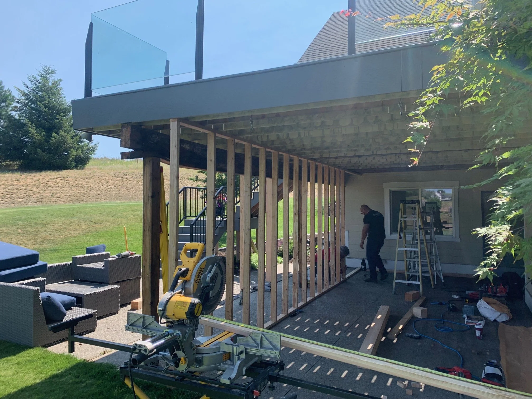 Construction workers building a deck on the back of a house, with tools and ladders present, and a glass railing on the upper level.