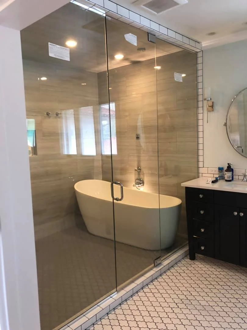 Bathroom with glass-enclosed shower area featuring a large white freestanding bathtub, a black vanity with dual sinks, and hexagonal tiled flooring.