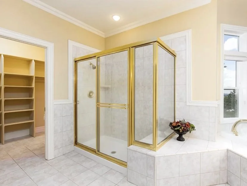 Bathroom with a glass shower enclosure with a gold frame, a built-in tub with a flower arrangement, and a doorway leading to a walk-in closet with shelves.