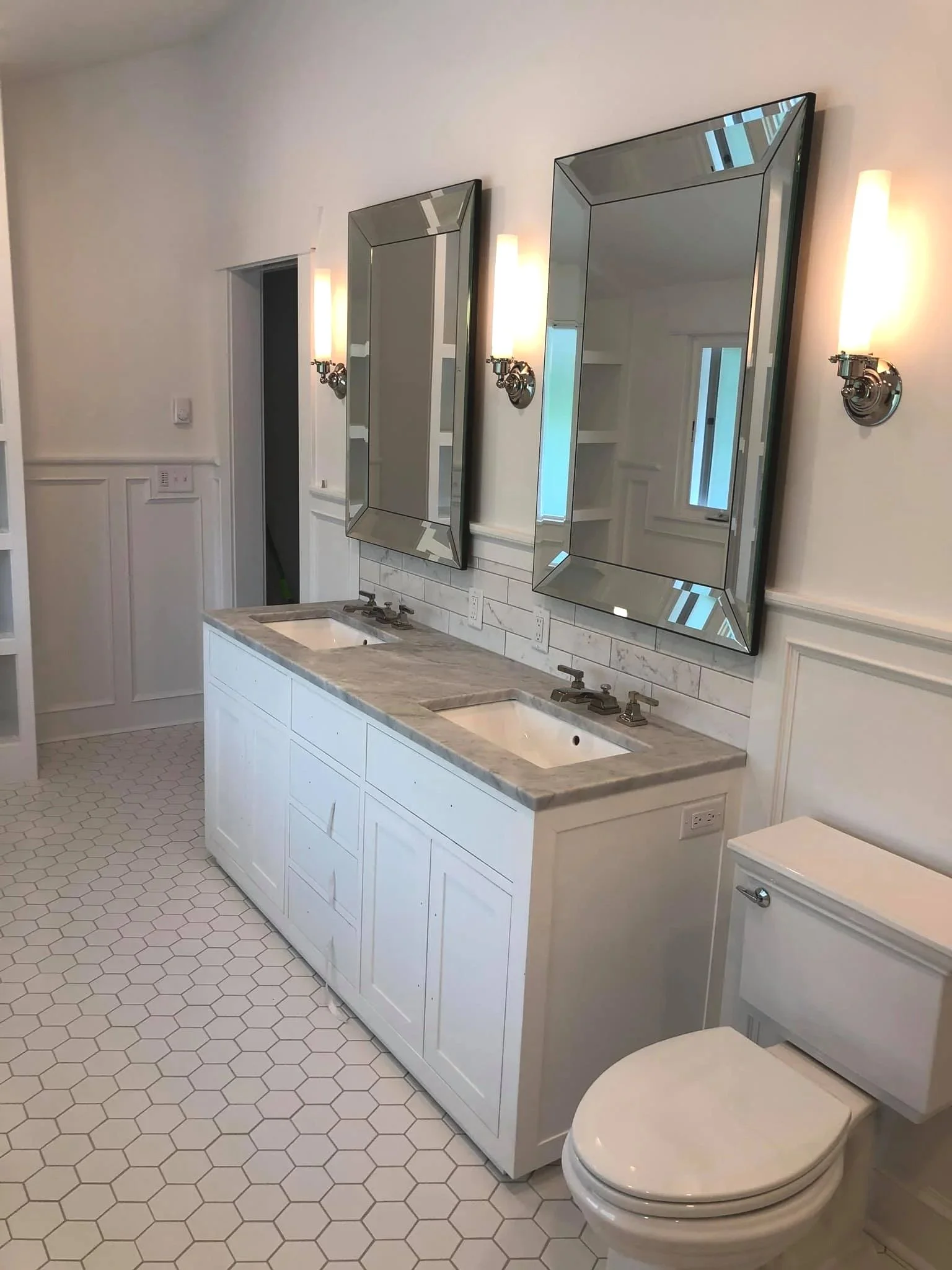 A bathroom with a double vanity, two mirrors, two wall sconces, and a toilet. The vanity has a marble countertop with two sinks and bronze fixtures. The floor is covered with hexagon tiles.