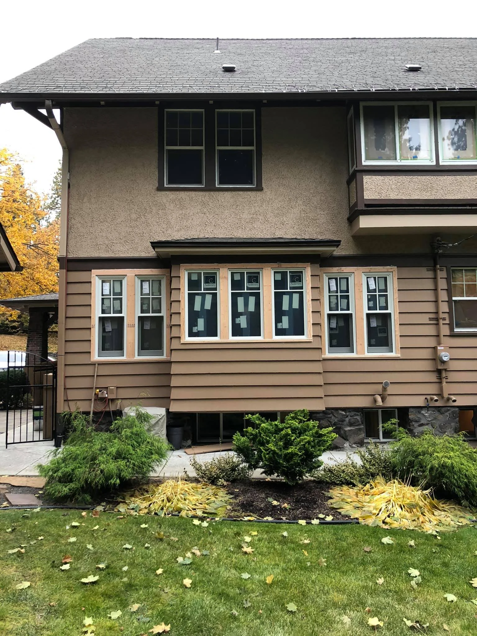House undergoing renovation with new windows installed on the lower level, front yard with green and yellow plants, and fallen leaves.