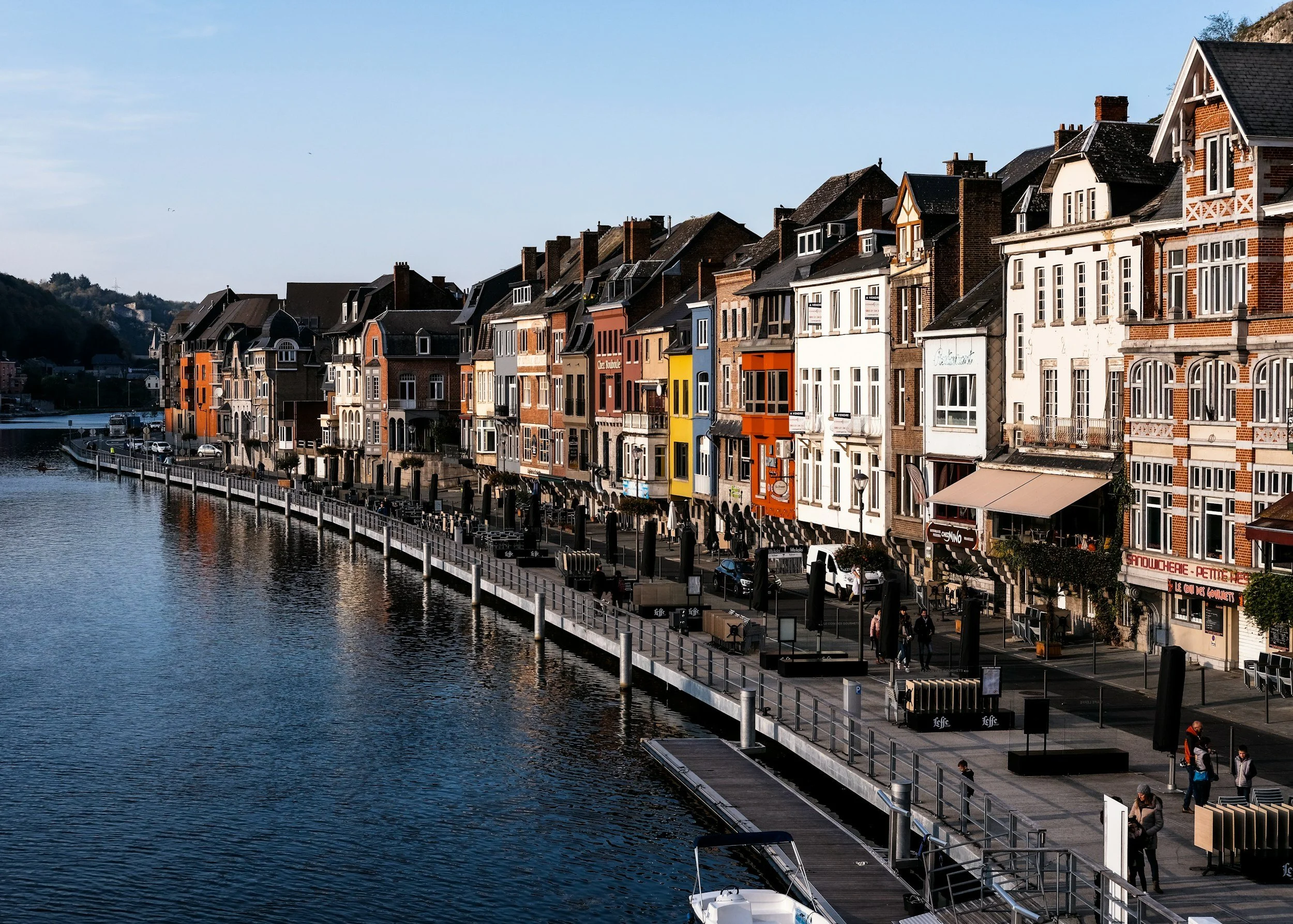Row of colorful houses along a waterfront promenade with boats and people walking, under a clear blue sky.