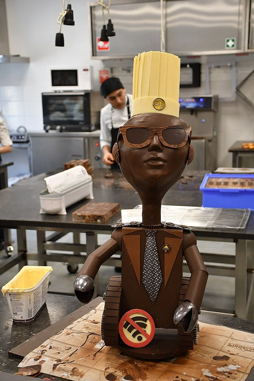 A chocolate sculpture of a person with a tall chef's hat, sunglasses, and a suit, displayed on a table in a bakery or kitchen setting with bakers working in the background. EUHOFA Congress.