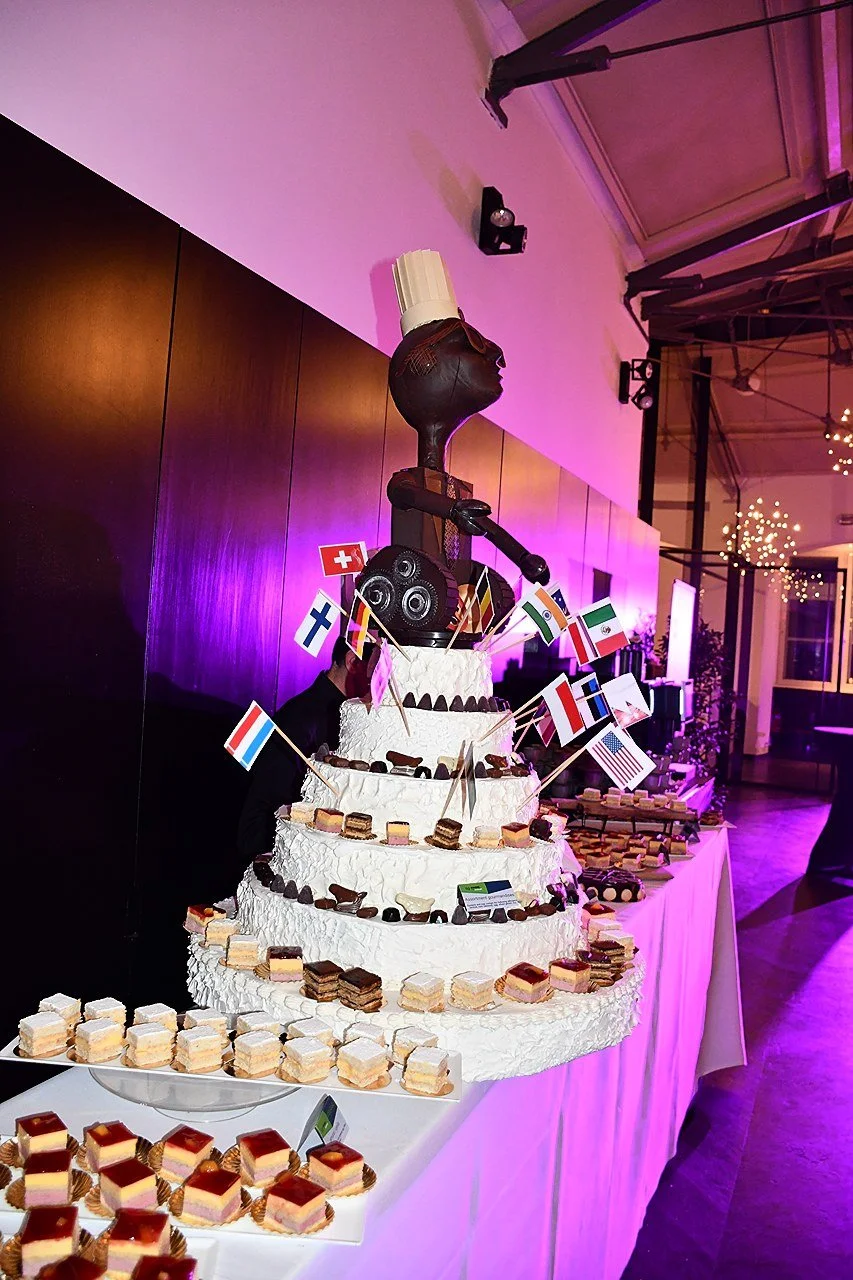 A large, tiered cake with a sculpture on top featuring a figure with sunglasses and a chef's hat, decorated with small international flags, in a banquet hall with purple lighting and a dessert table at EUHOFA Congress Belgium 2026.