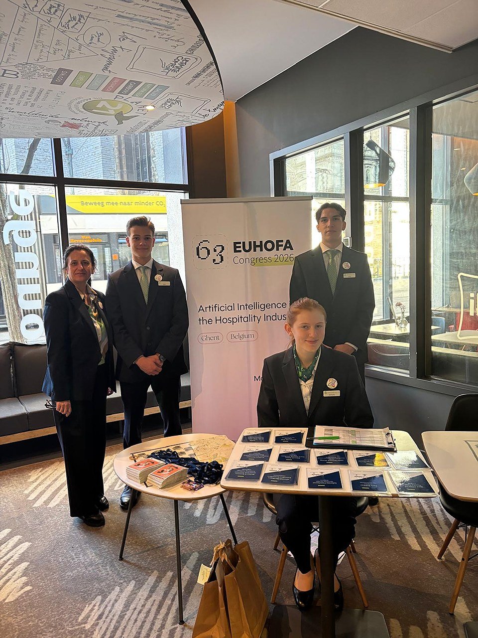 Group of four young students at a conference booth for EUHOFA Congress 2026, focusing on artificial intelligence in the hospitality industry, with promotional materials and banners.