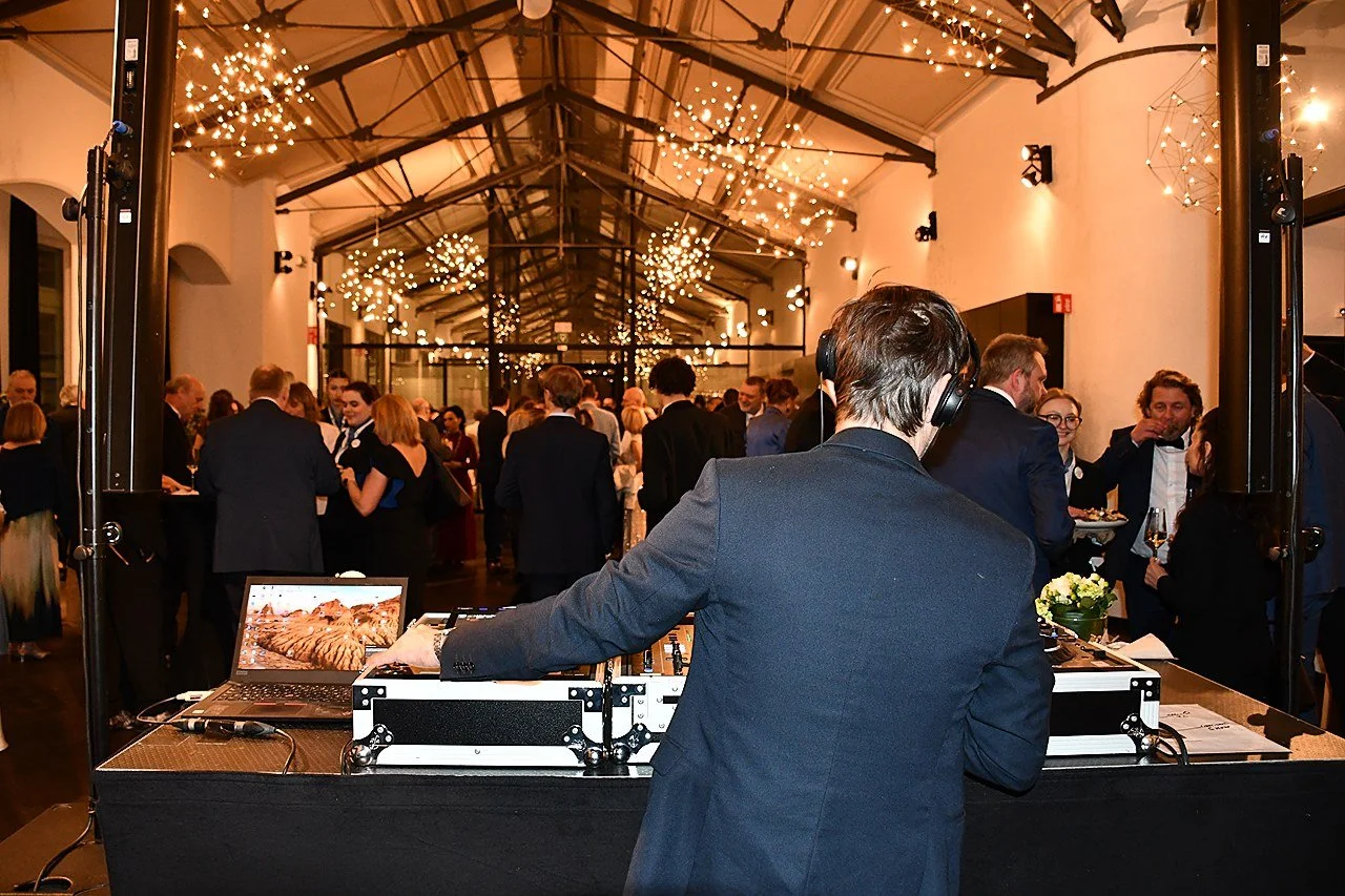 A DJ wearing headphones performs at a crowded formal event with members in tuxedos and evening gowns, under a ceiling decorated with string lights at EUHOFA Congress.