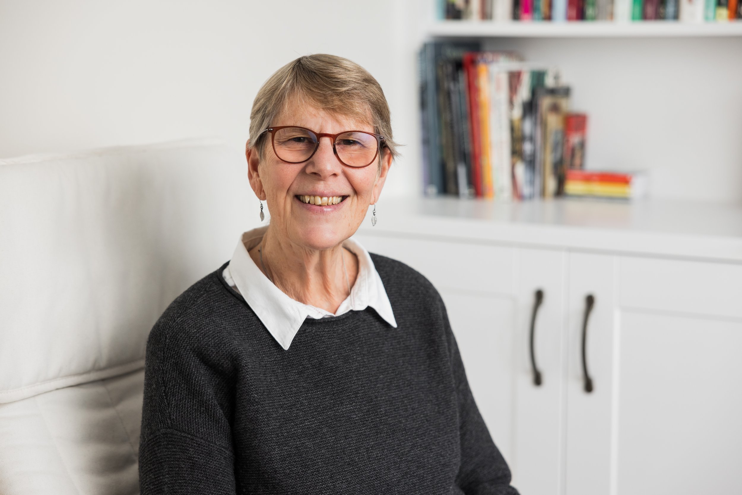 An image of Wendy Durell with short hair, glasses, and earrings, smiling while sitting on a white couch with a bookshelf in the background.