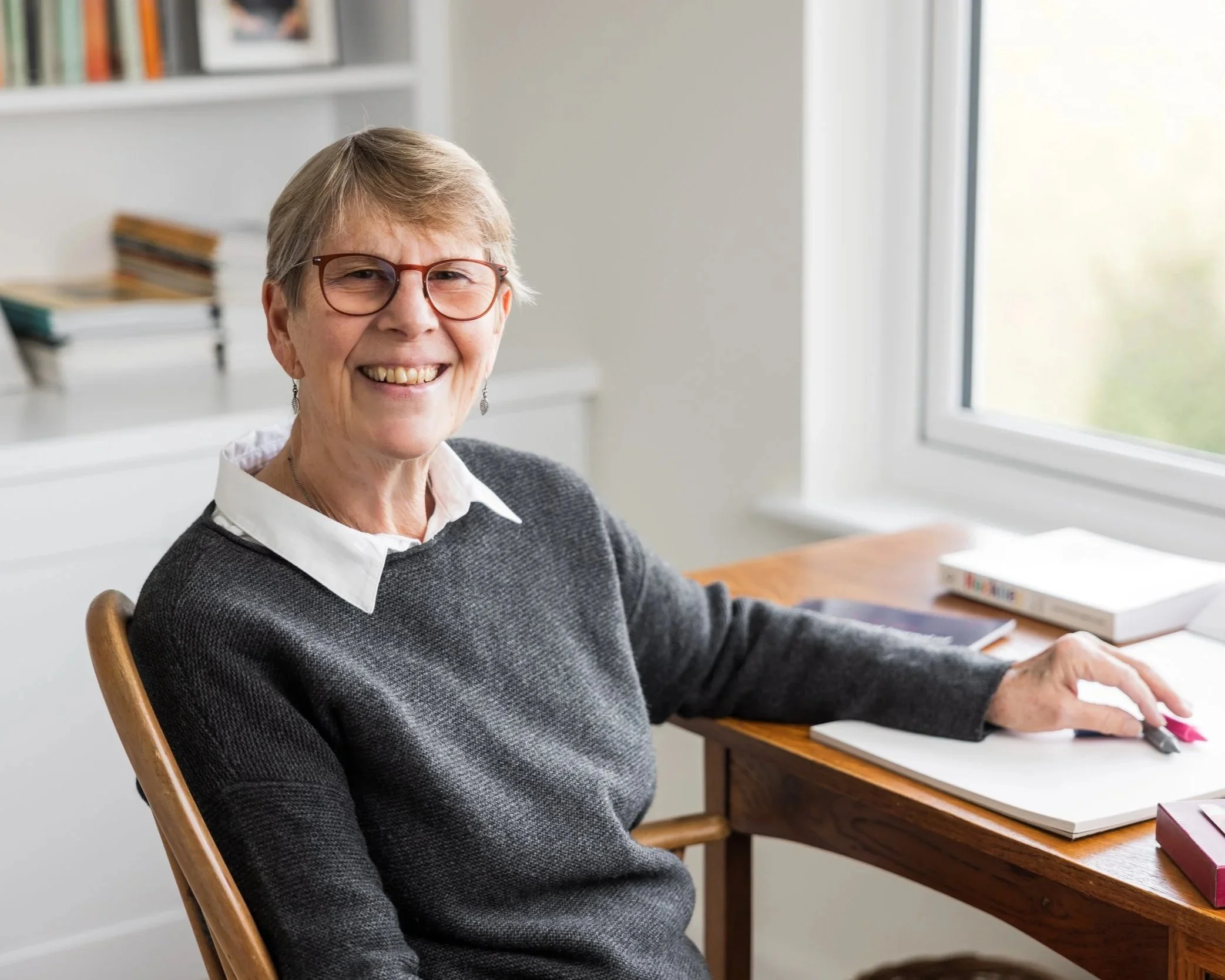 Smiling photo of Wendy Durell with short hair and glasses sitting at a wooden desk near a window in a bright room, wearing a white shirt and a dark sweater.