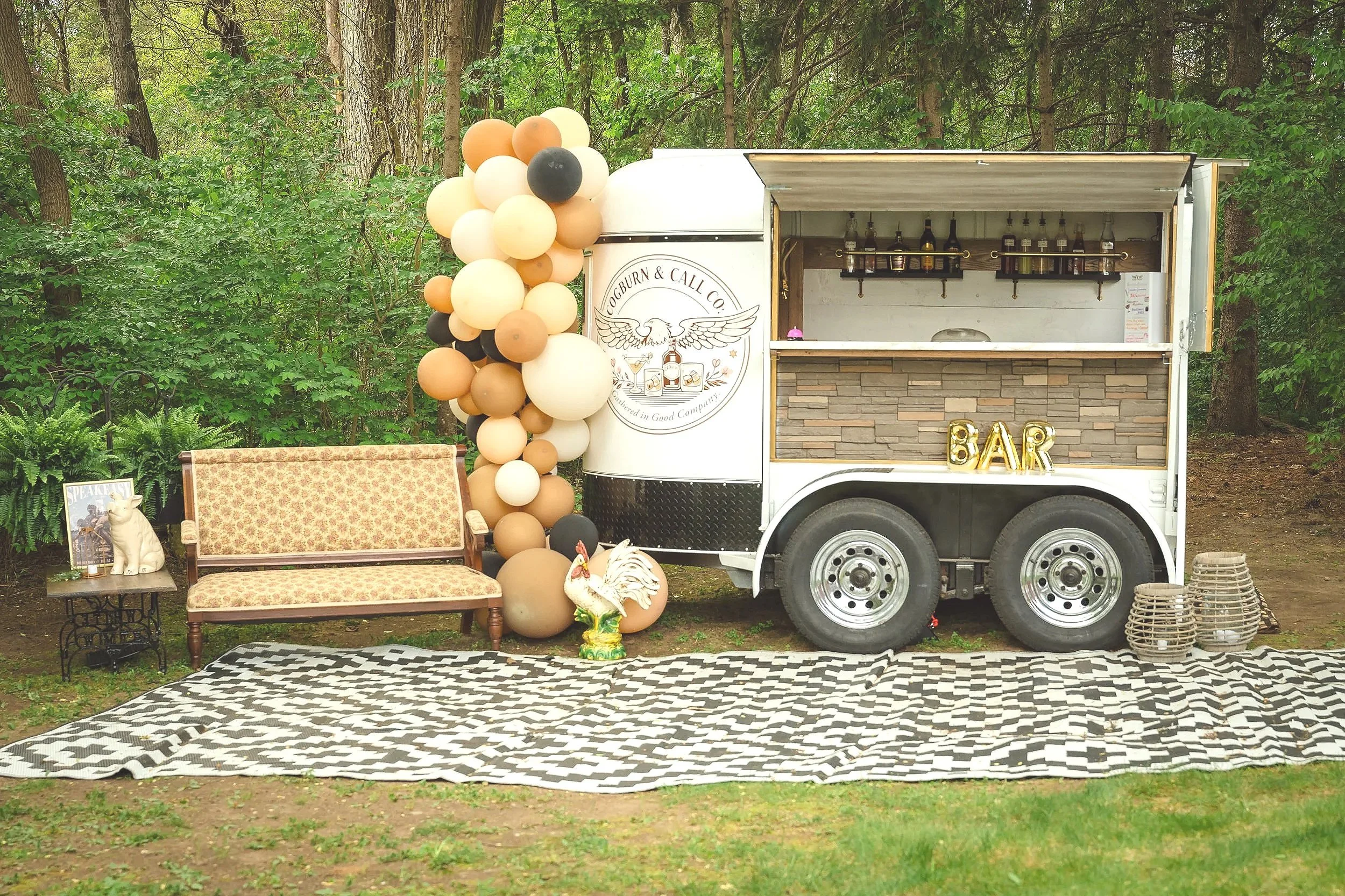 A mobile bar trailer decorated with a balloon garland, with a sign that says 'BAR' and a logo that reads 'Oakbourn & Call Co.' featuring a bird with outstretched wings. To the left, there is a vintage-style sofa, a small table with a dog sculpture, and various decorative items. The setting is outdoors in a lush, wooded area, with a patterned rug on the ground.