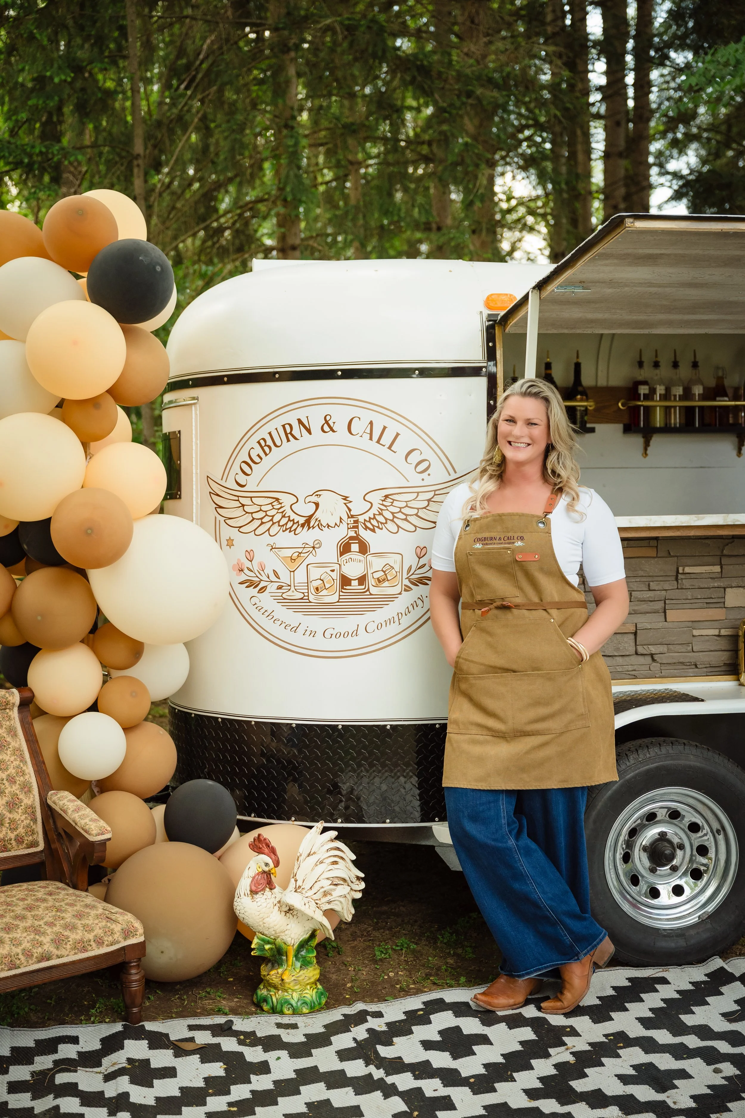 A woman with blonde hair, wearing a white shirt, brown apron, and blue jeans, standing in front of a vintage food truck with a logo that reads 'Cobburn & Call Co.' surrounded by balloons in shades of white, gray, black, and tan. The truck is parked outdoors with a background of trees. In front of her, there is a decorative chicken statue, a floral armchair, and a black-and-white patterned rug.