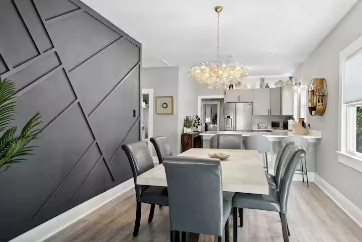 Modern dining room with gray chairs around a white table, dark accent wall with geometric patterns, and a bright kitchen with white cabinets and stainless steel appliances.