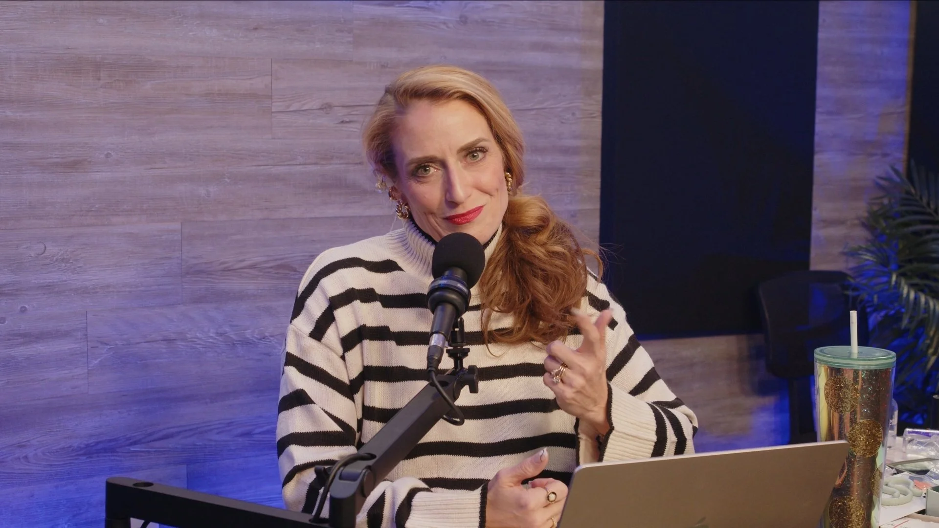 Woman with long red hair and earrings speaking into a microphone in a recording studio, wearing a black and white striped sweater, with a drink with a straw and a laptop in front of her.