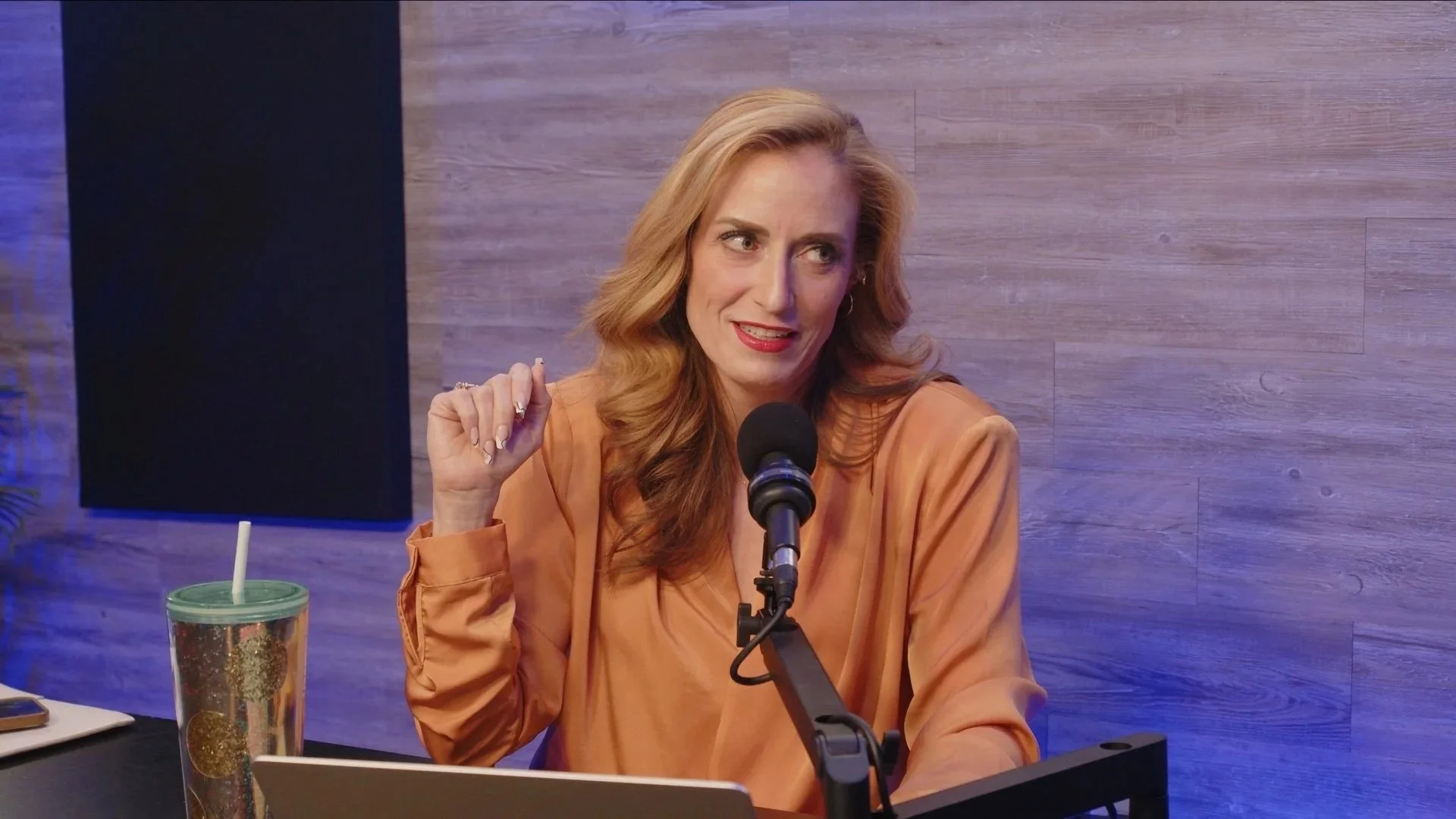 A woman with long, wavy red hair sits at a radio or podcast studio table, speaking into a microphone. She is wearing a light brown or tan blouse, with a glass of iced drink with a straw in front of her. The background features a wooden wall and purple lighting.