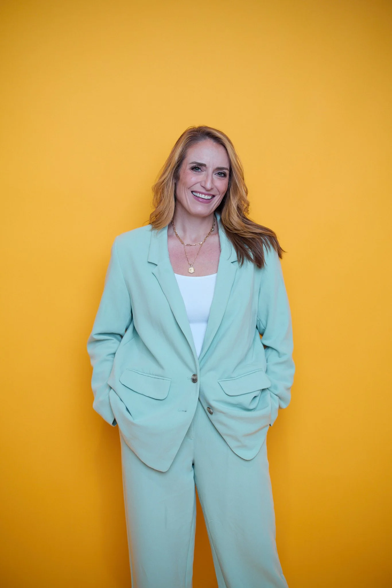 A woman with red hair, wearing a pastel blue suit and layered necklaces, is smiling at the camera against a bright yellow background.