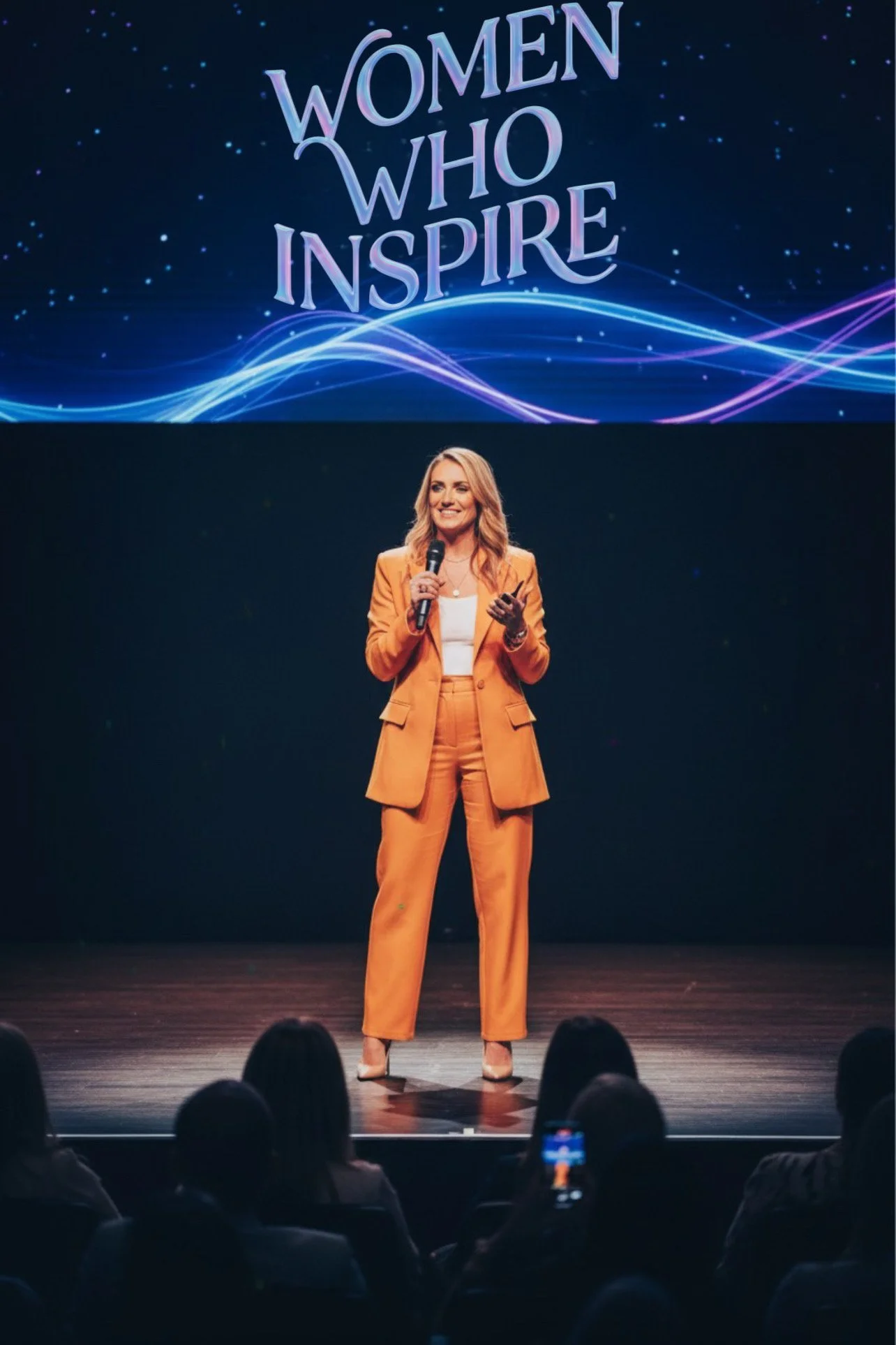 A woman in an orange suit speaking on stage with a microphone, with a large screen behind her displaying the words 'Women Who Inspire' and colorful graphical lines.