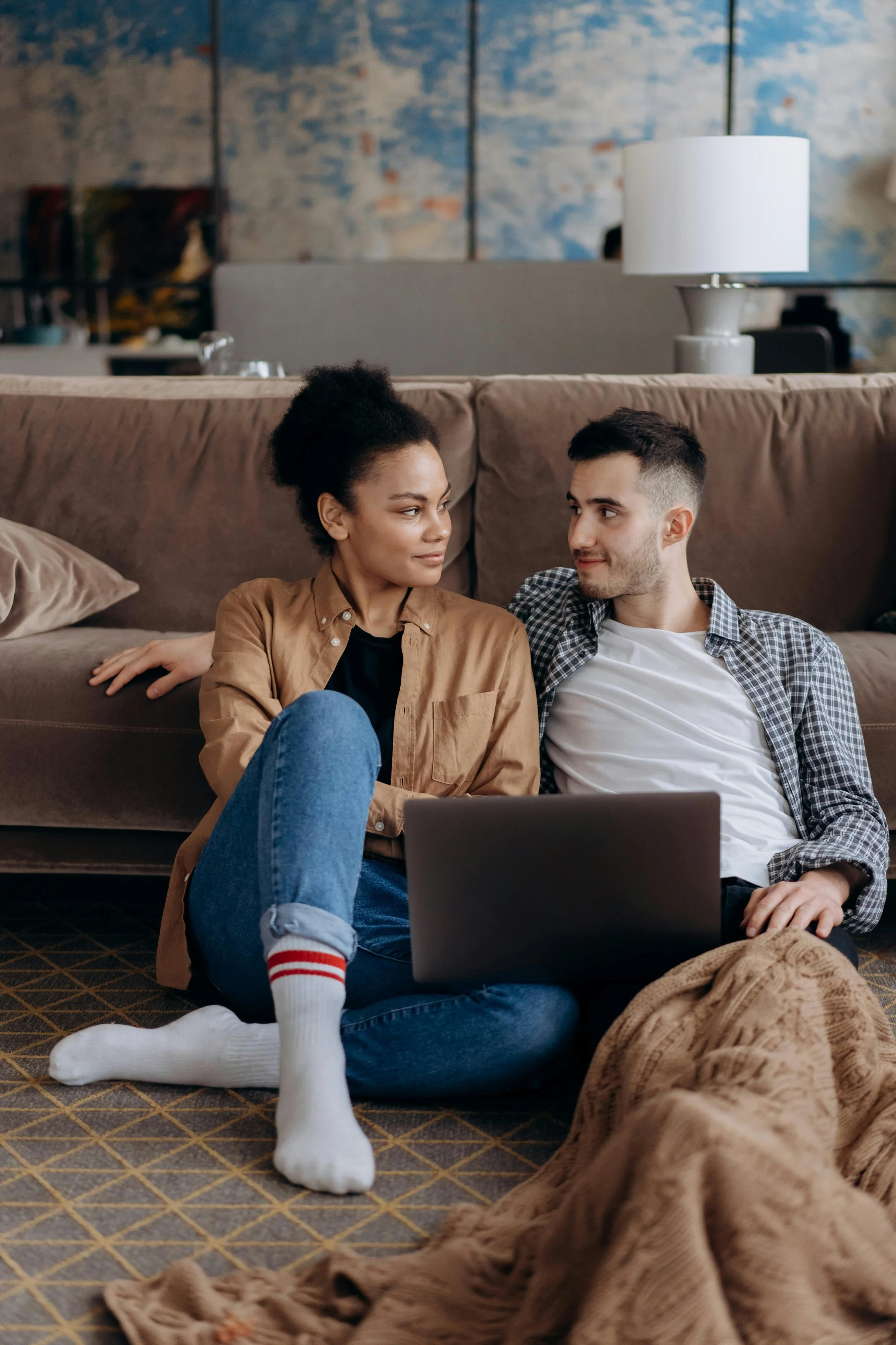 A man and woman sitting on the floor of a living room, leaning against a sofa, looking at each other. The woman has curly hair in a bun and is wearing a brown shirt and jeans. The man has short dark hair and is wearing a checkered shirt over a white T-shirt. There is a laptop on their lap, and a blanket is on the floor in front of them. The background shows a sofa, a lamp, and a patterned wall.
