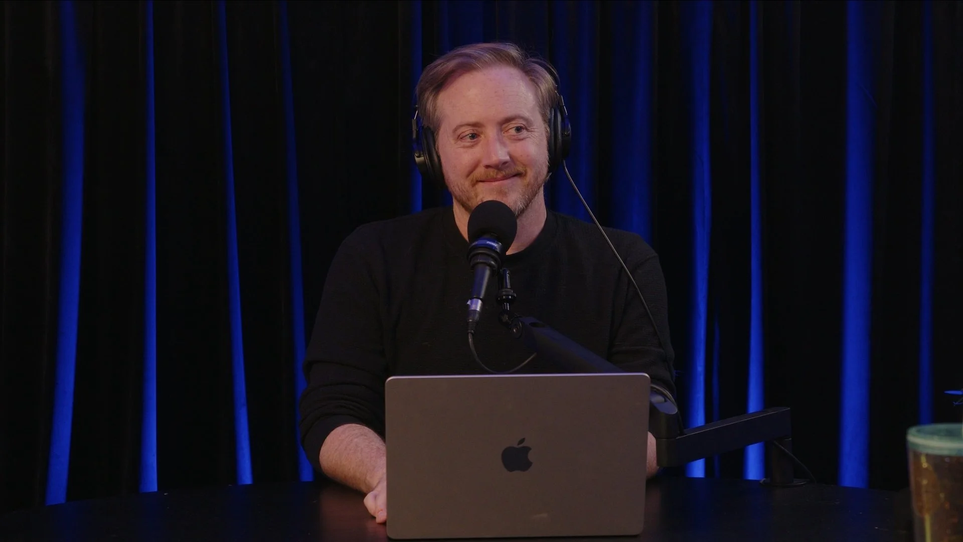 A man with reddish hair and a beard, wearing a black sweater and headphones, sits at a table with a silver MacBook, speaking into a microphone against a black curtain with blue vertical stripes.