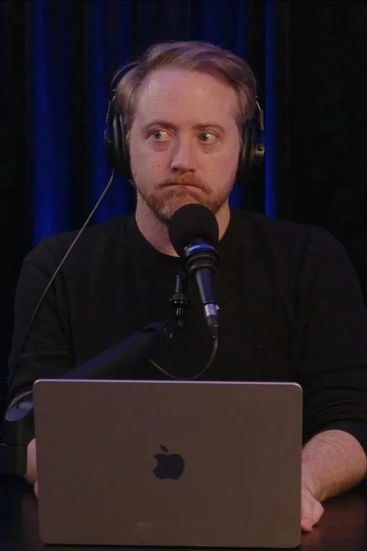 A man with red hair and a beard, wearing headphones, sitting in front of a microphone and a silver MacBook laptop, on a dark background.