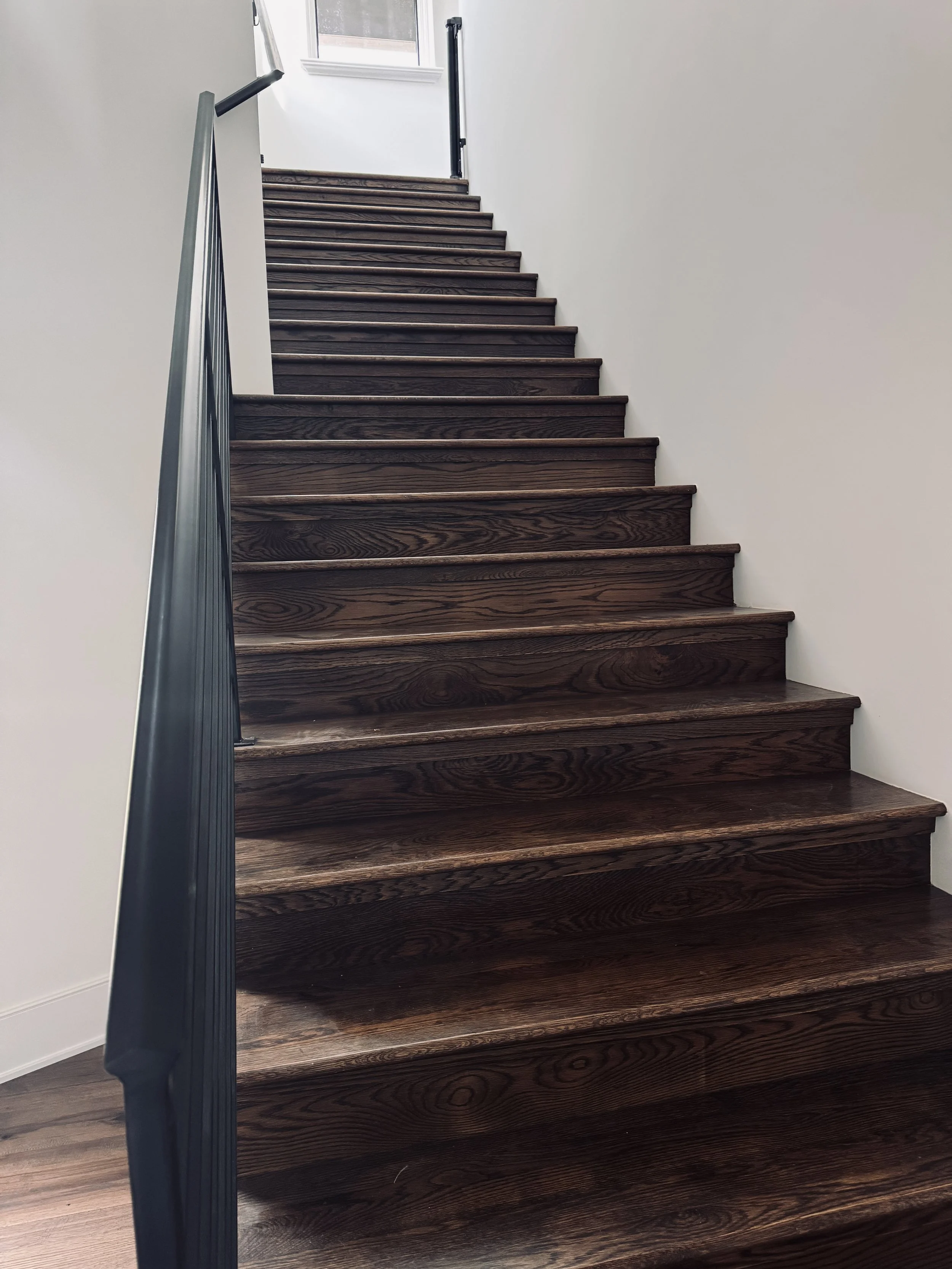 Interior view of a staircase with dark wooden steps, black railing, white walls, and a window at the top of the stairs.
