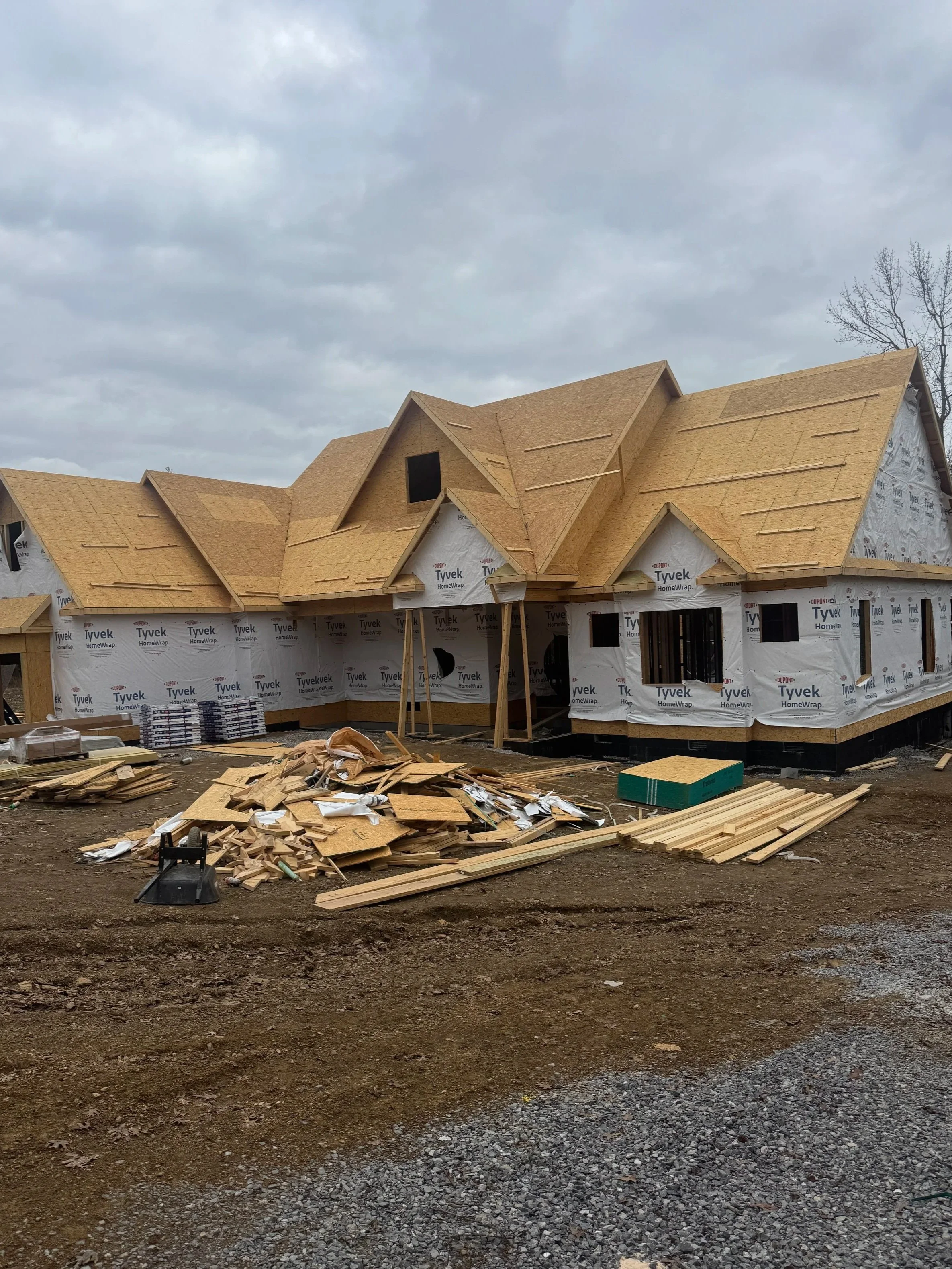 Under construction house with wooden roof framing and construction materials scattered on the ground, cloudy sky in the background.