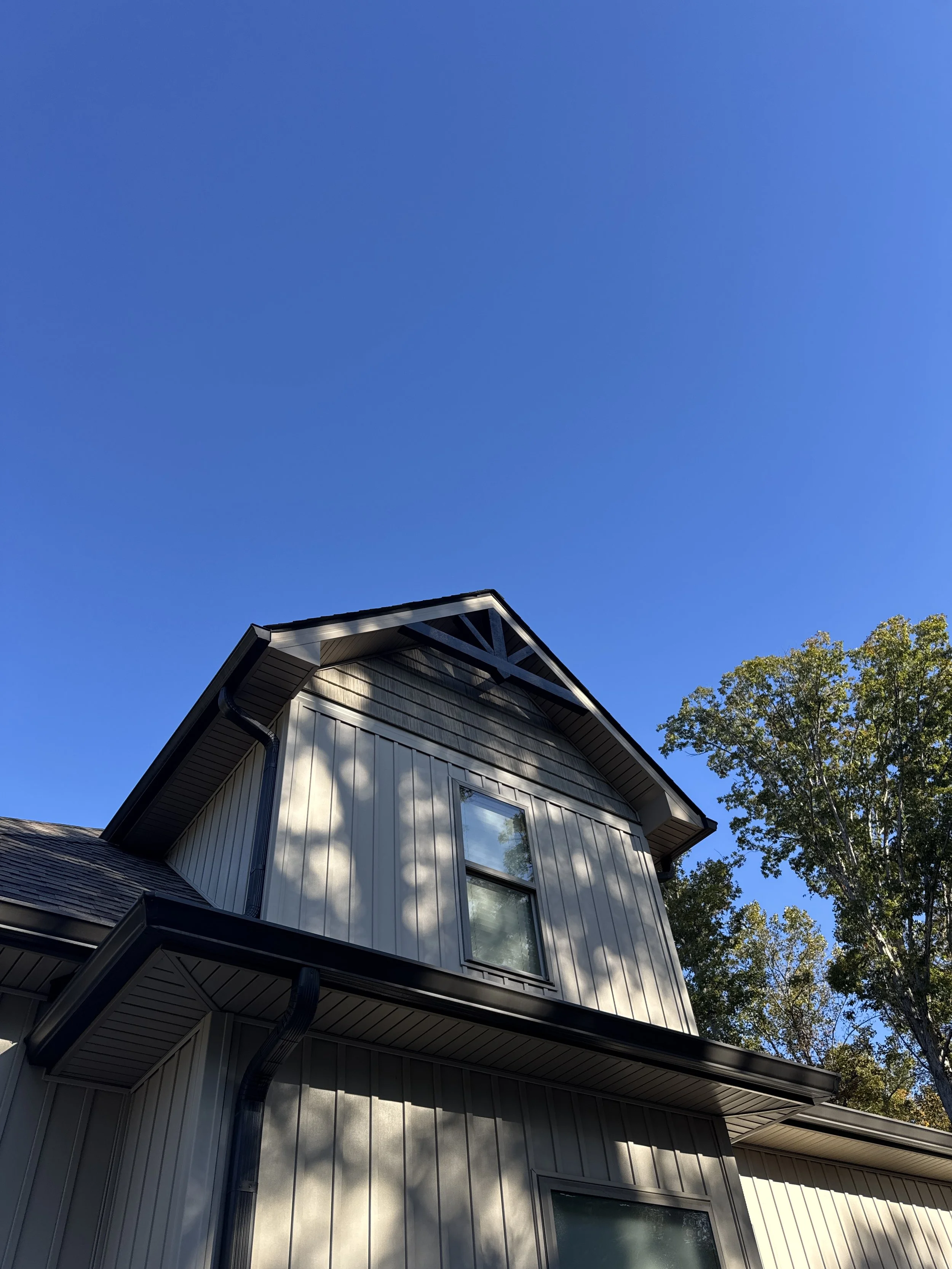 A residential house with white siding and large windows, surrounded by trees; the roof has a prominent gable, and the sky is clear and blue.