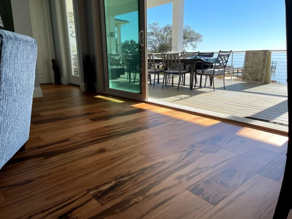 Living room showing hardwood floors opening to a deck with outdoor dining furniture and a railing overlooking a body of water and trees.