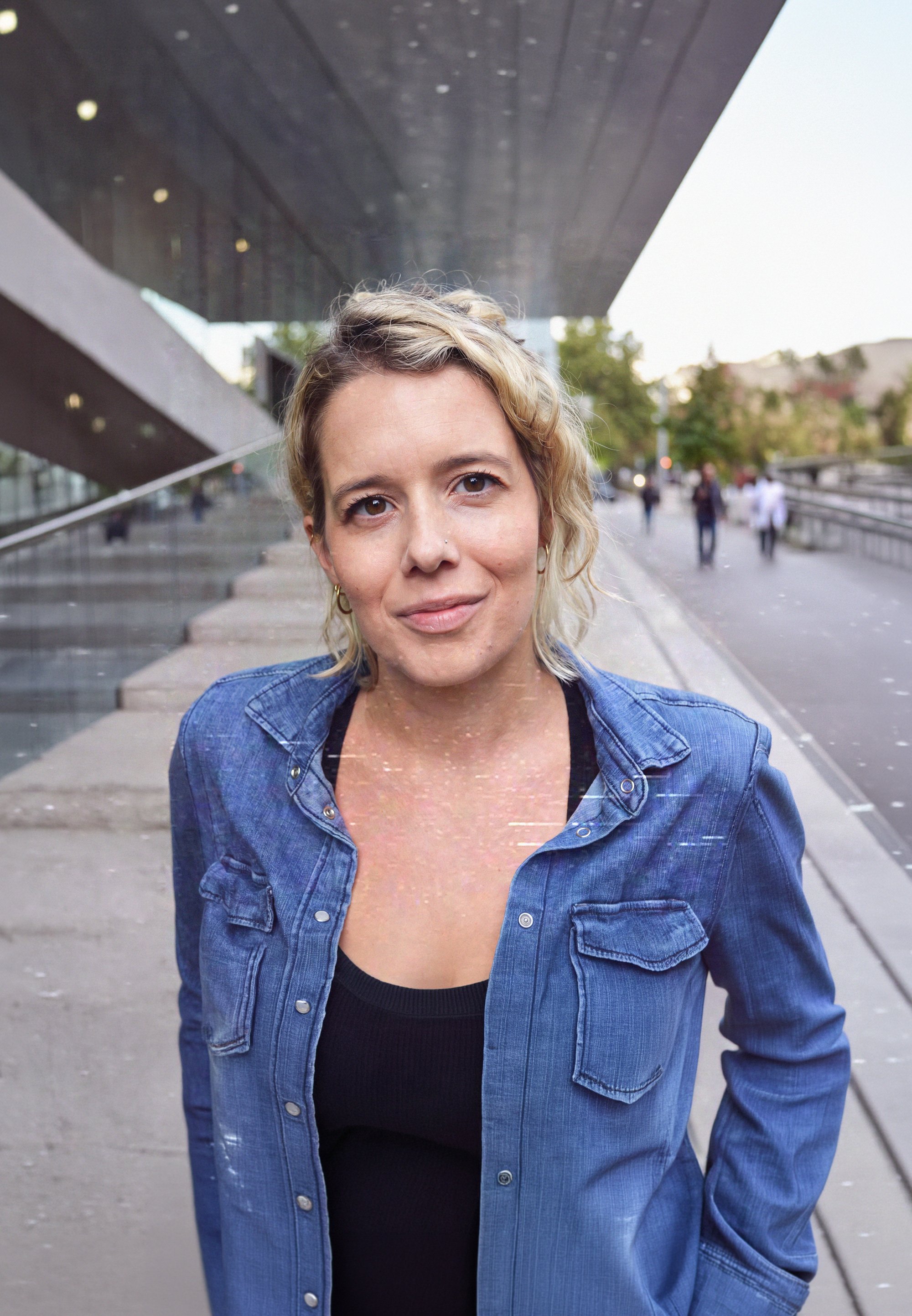 A woman with blonde hair, wearing a black top and blue denim jacket, standing outdoors on a city sidewalk near a modern building with large overhangs and glass windows, with people walking in the background.