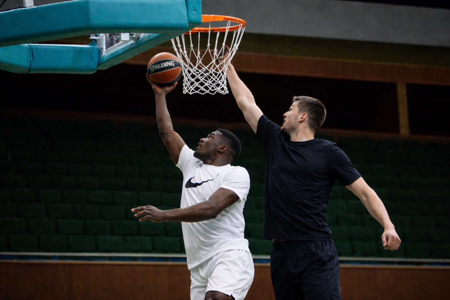 Two men playing basketball indoors, one in a white shirt and shorts attempting a shot near the hoop, the other in a black shirt jumping to defend. The players are participating in a CLA (Constraints Led Approach) supported by Ecological Dynamics.
