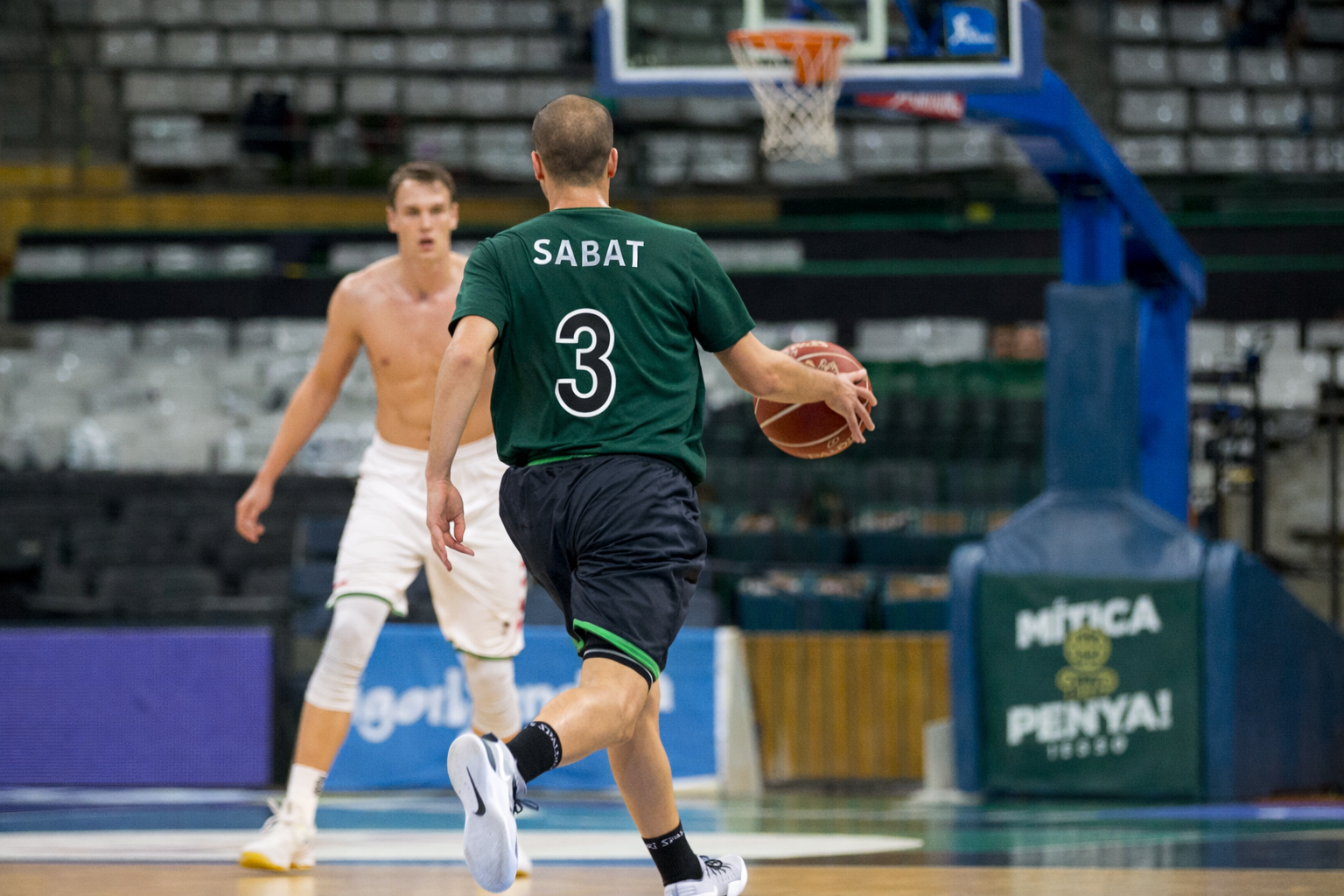 Two basketball players on an indoor court, one in a green jersey holding a basketball, while the other player, shirtless and wearing white shorts, looks on. There is a basketball hoop and backboard in the background.