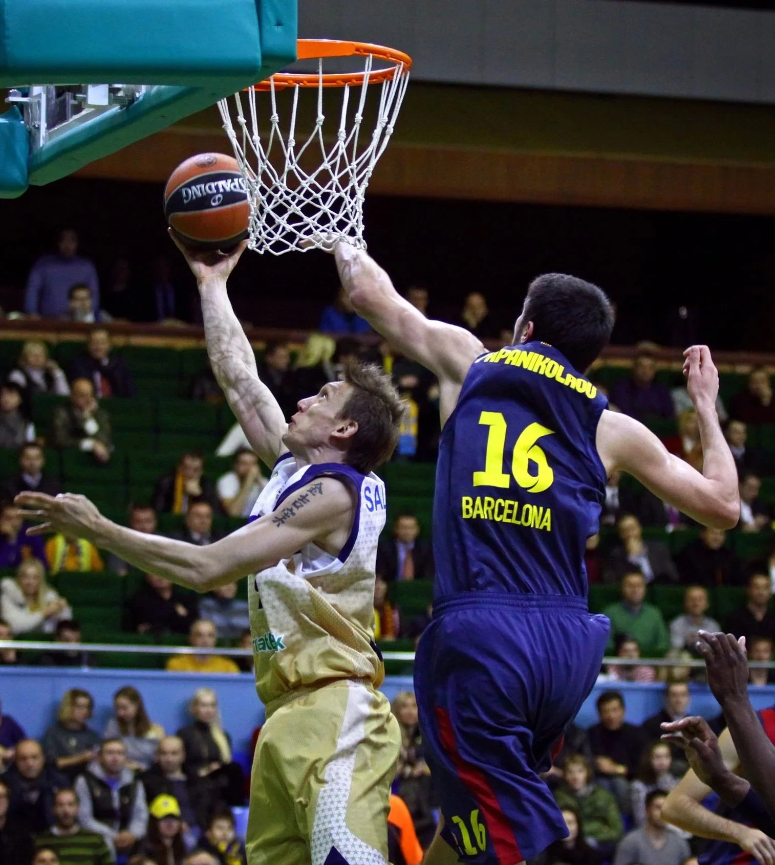 A basketball player in a white and gold jersey slam-dunks the ball into the hoop while an opposing player in a blue and yellow jersey attempts to block the shot during a Constraints Led Approach basketball workout.