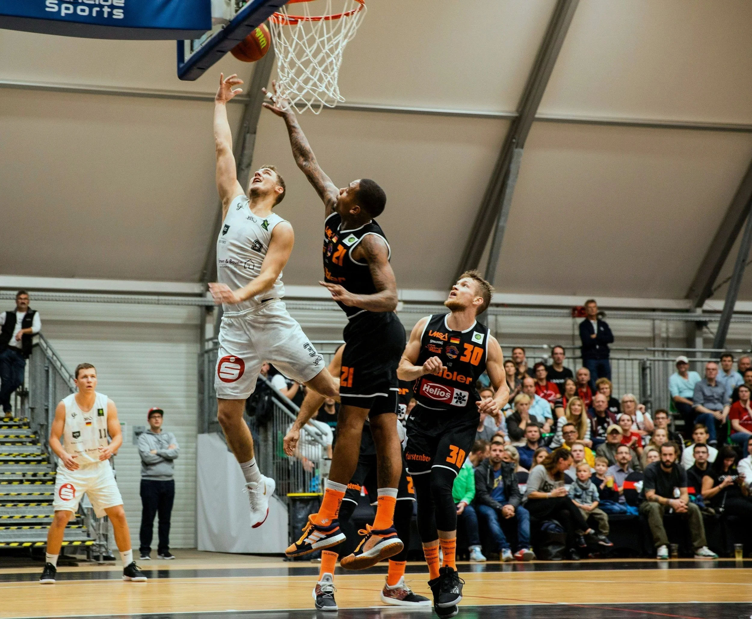 Basketball players in mid-air attempting a shot or block during a game, with spectators watching from the stands. The players are participating in a CLA (Constraints Led Approach) supported by an Ecological Dynamics framework.
