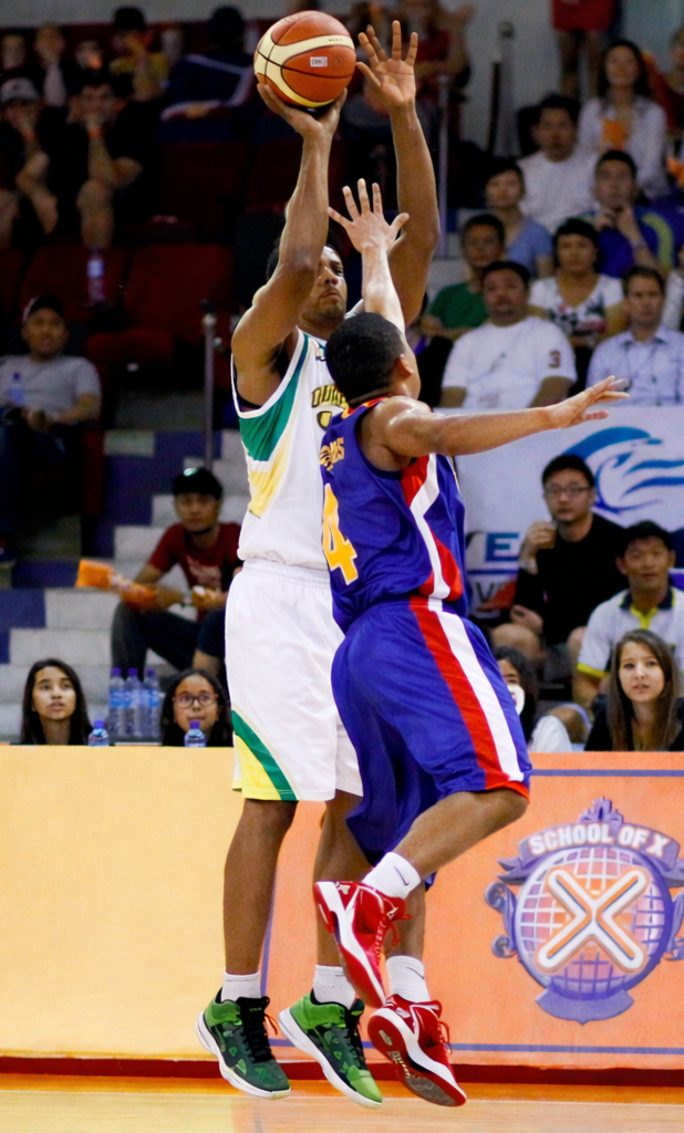 Two basketball players in mid-air during a Constraints Led Approach basketball game. One player, in a white and green uniform, is shooting the basketball. The other player, in a blue and red uniform, is defending.