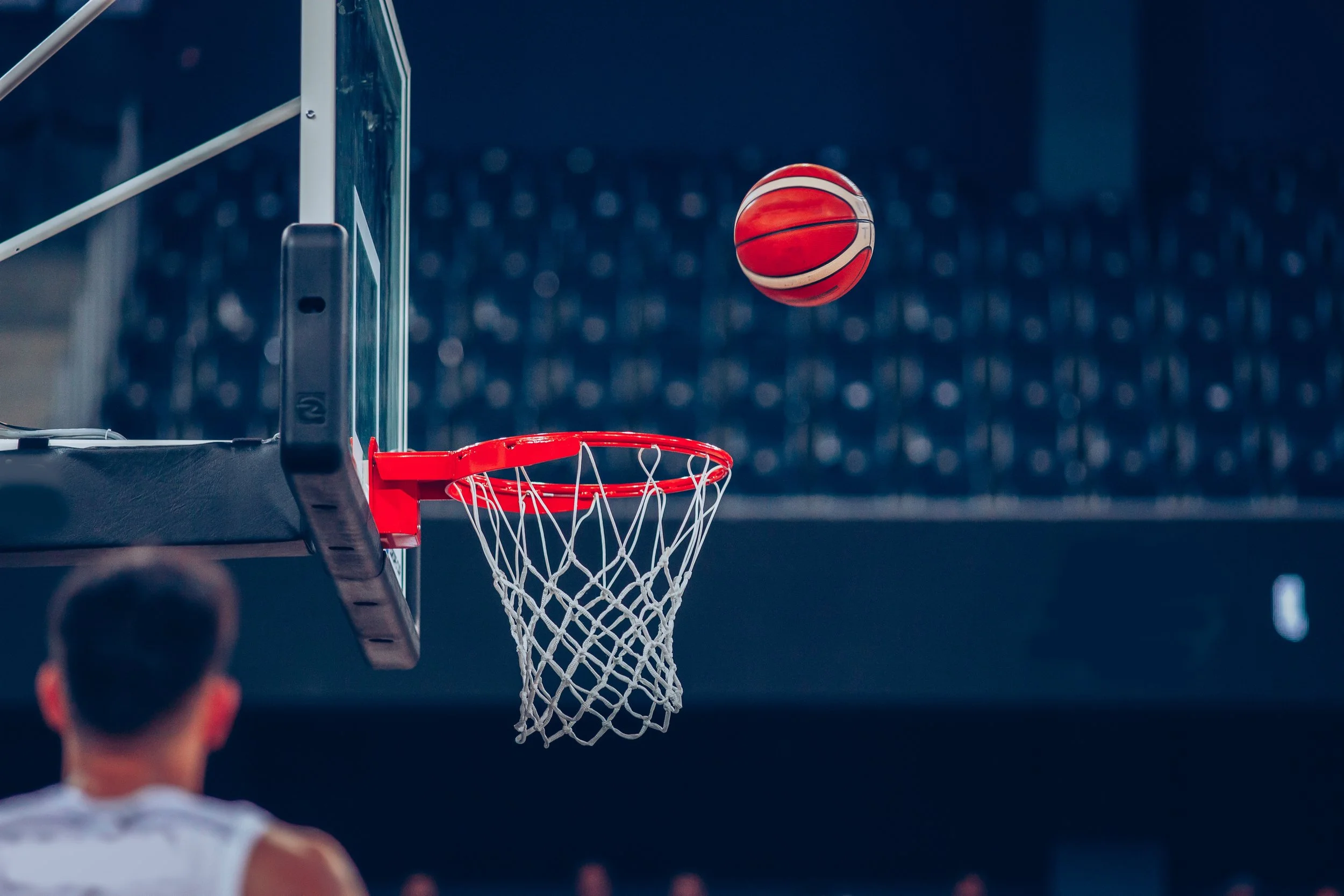 A basketball is seen flying towards the hoop on an indoor basketball court, with a player watching in the foreground. The player is participating in a CLA (Constraints Led Approach) supported by an Ecological Dynamics framework.