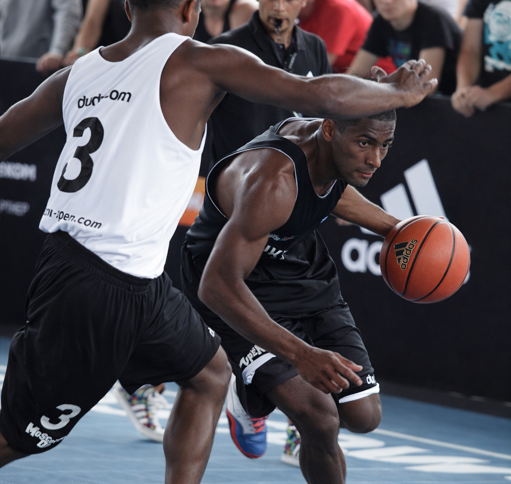 Two basketball players, one in black and one in white, compete during a game on an indoor court with spectators in the background. The players are participating in a CLA (Constraints Led Approach) supported by an Ecological Dynamics framework.