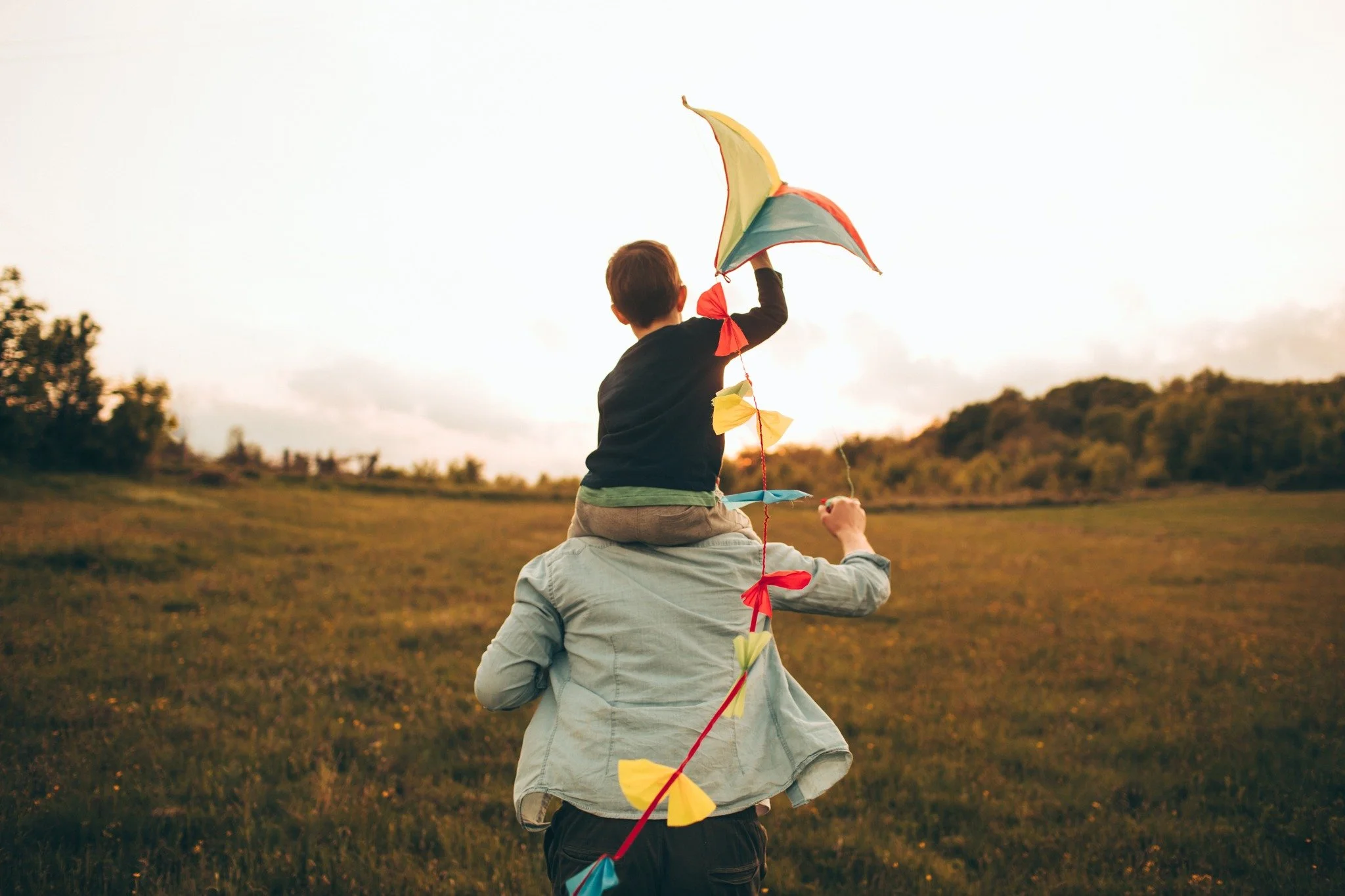 A child sitting on an adult's shoulders holding a colorful kite outdoors during sunset.