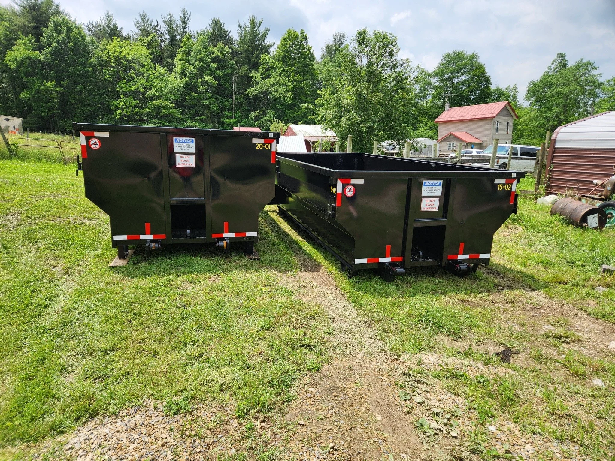 Two large black dumpsters with warning labels on a grassy area, surrounded by trees and rural buildings and structures in the background.
